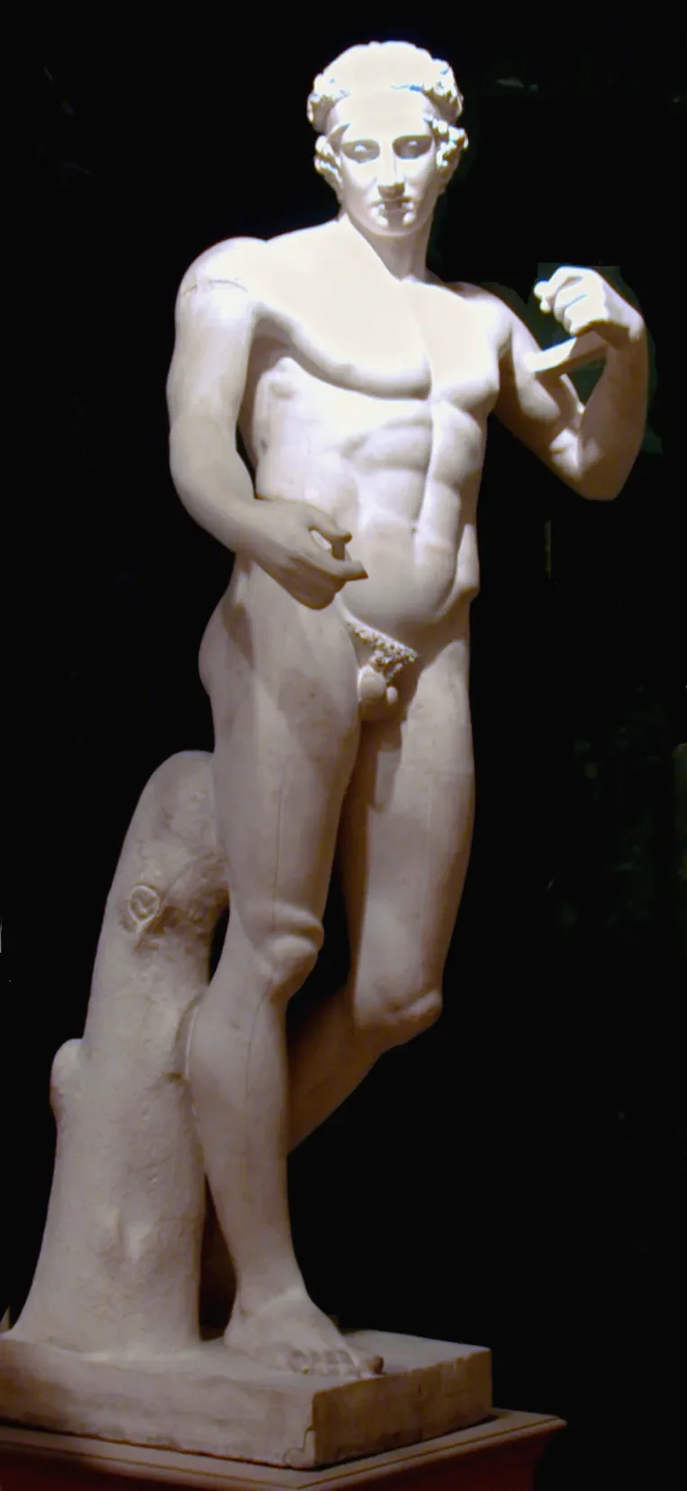 Escultura clásica de un hombre desnudo con barba y cabello rizado, en pose de fuerza, de pie sobre una base de piedra negra.