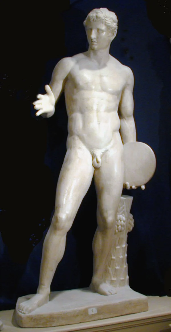 Escultura clásica de un hombre desnudo con pintura blanca, con un escudo en la mano derecha y mirando hacia la izquierda.