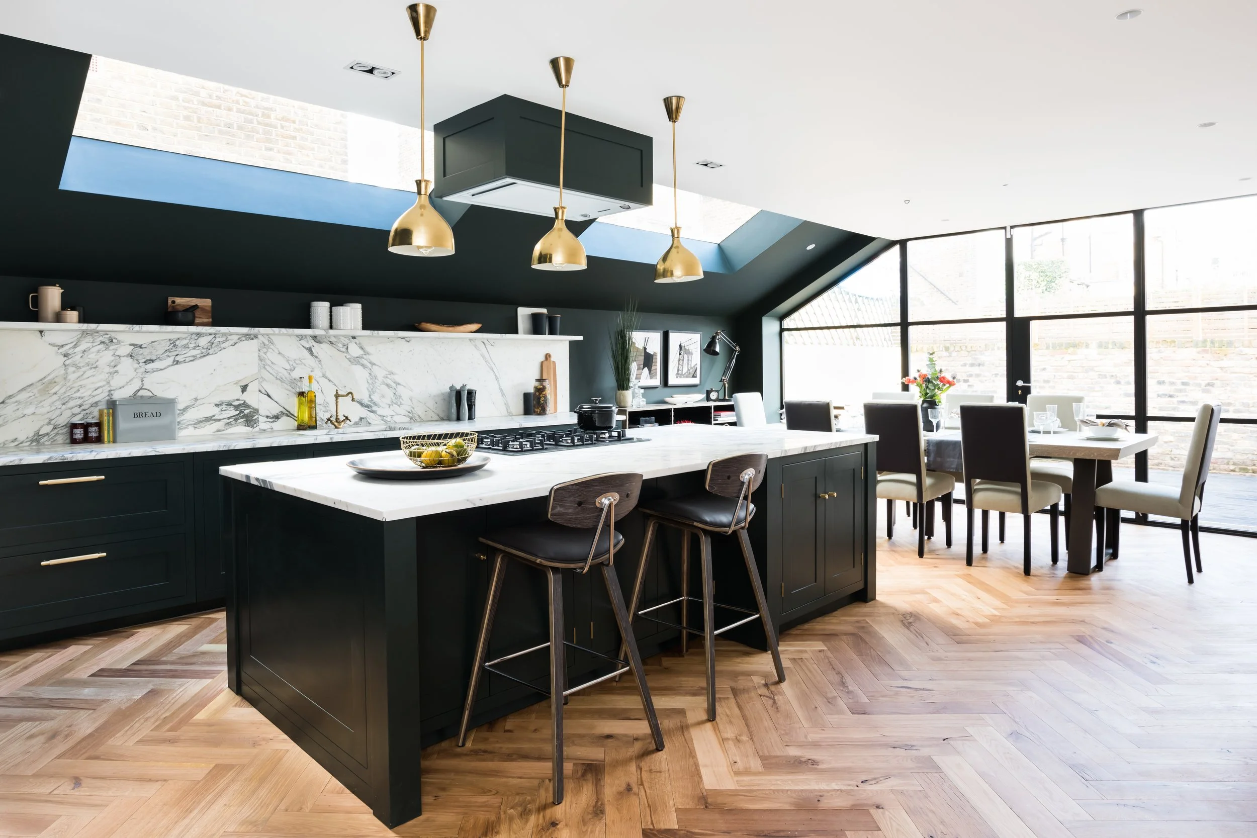 Cocina moderna con isla negra, encimeras de mármol y comedor con sillas blancas, ventanas grandes y suelo de madera