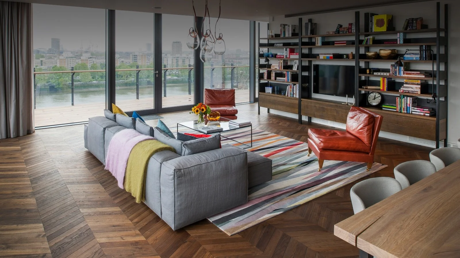 Sala de estar moderna con sofá gris, sillones de cuero rojo, estanterías negras con libros y una televisión, vista a un balcón con vista a un río y ciudad