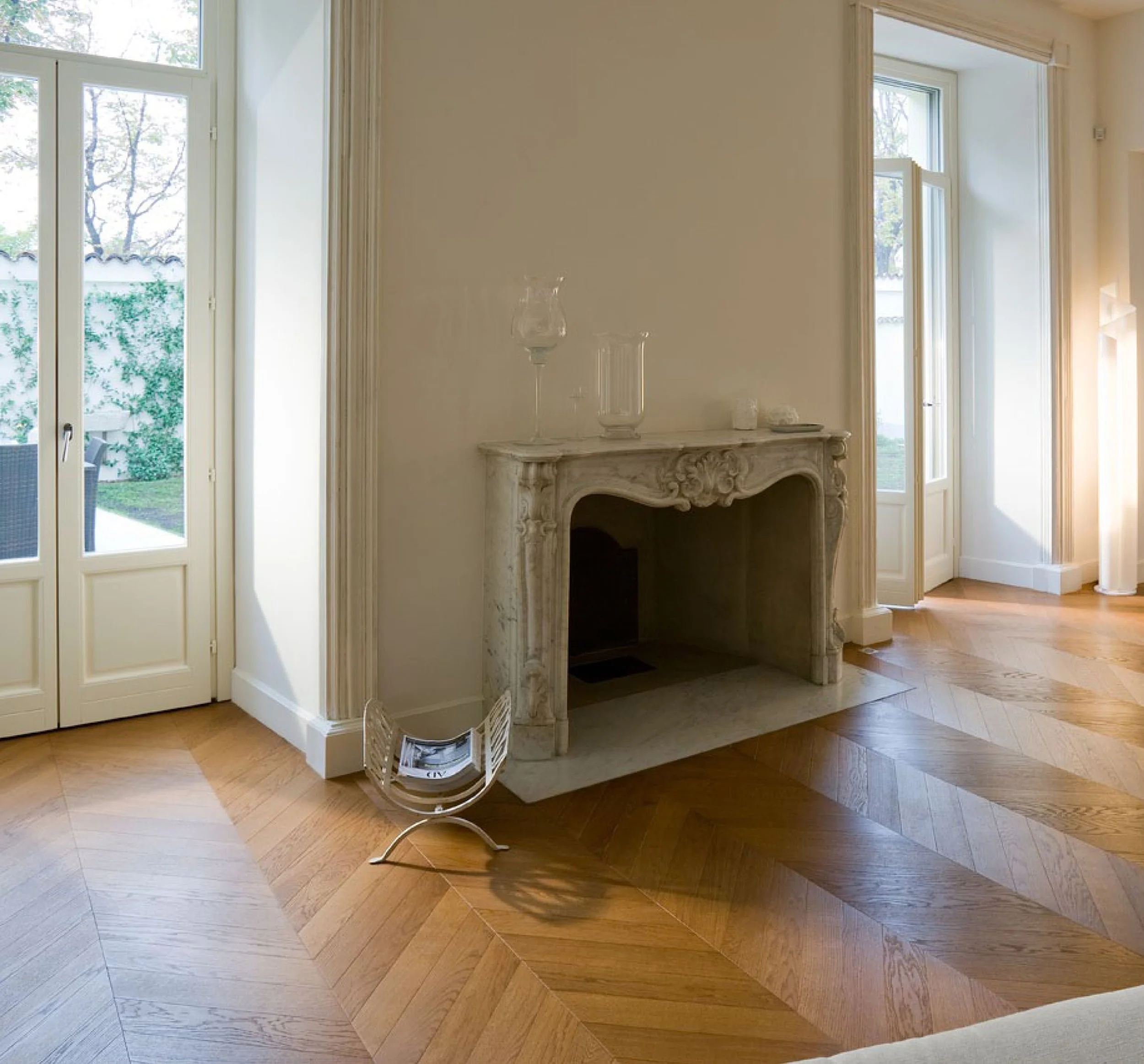 Sala con chimenea de mármol decorativa, puertas con cristaleras que dan al jardín, y suelo de parquet de madera en patrón diagonal.