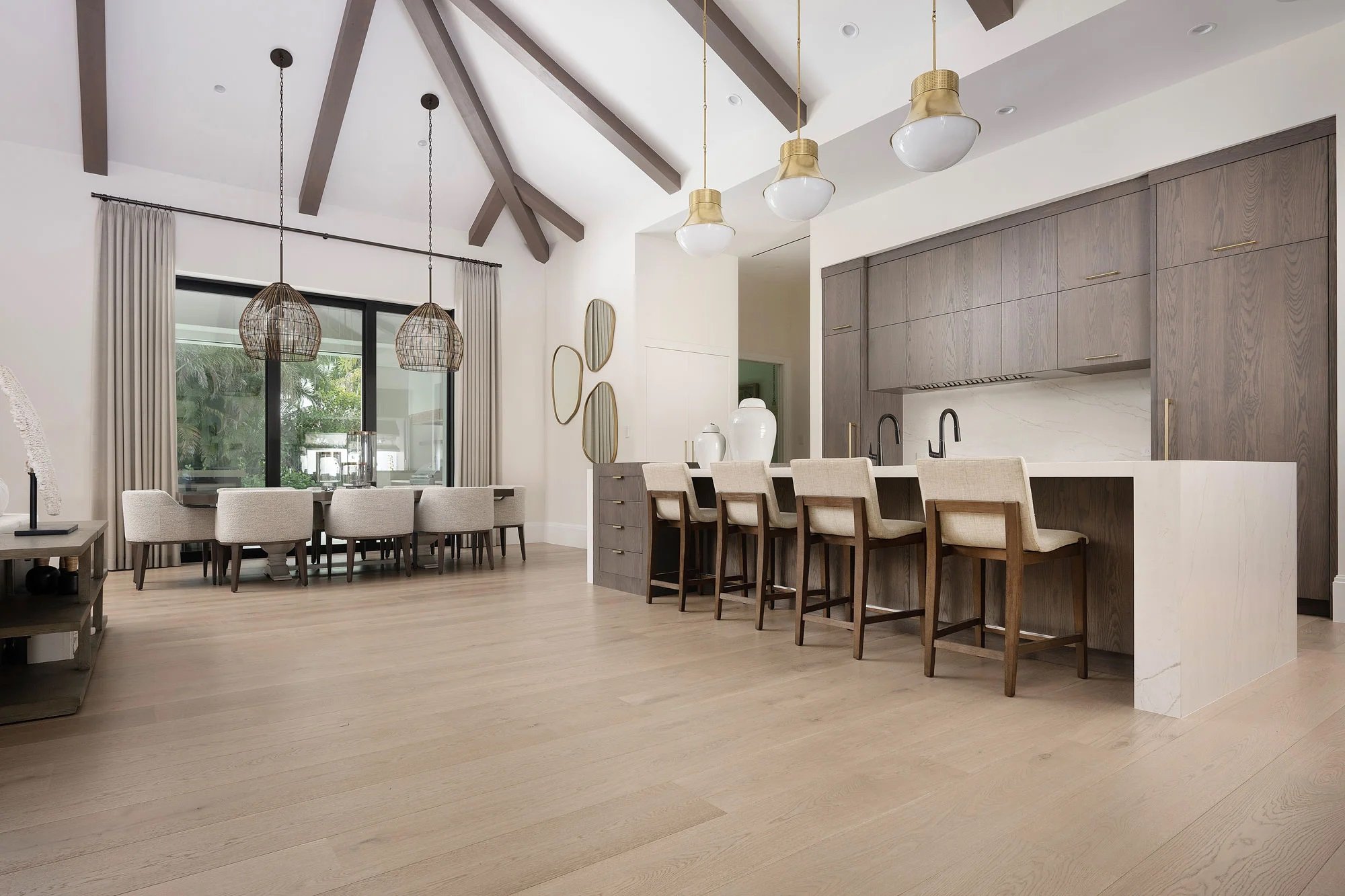 suelos de madera en cocina moderna con barra de desayuno, seis sillas, y comedor con ocho sillas junto a grandes ventanas con cortinas beige. Techos altos con vigas de madera expuestas.