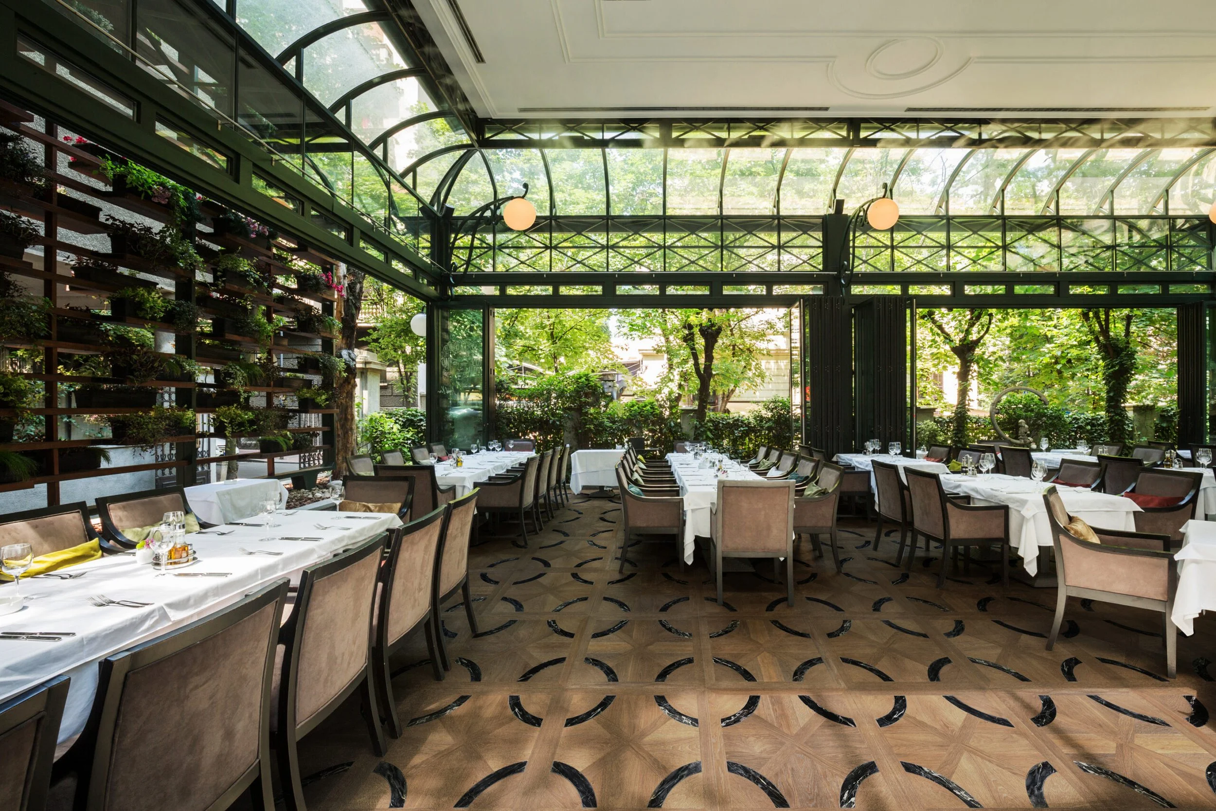 Restaurante elegante con mesas vestidas con manteles blancos y utensilios, rodeado de plantas y árboles, con paredes de cristal y un techo de vidrio curvo que permite la entrada de luz natural.