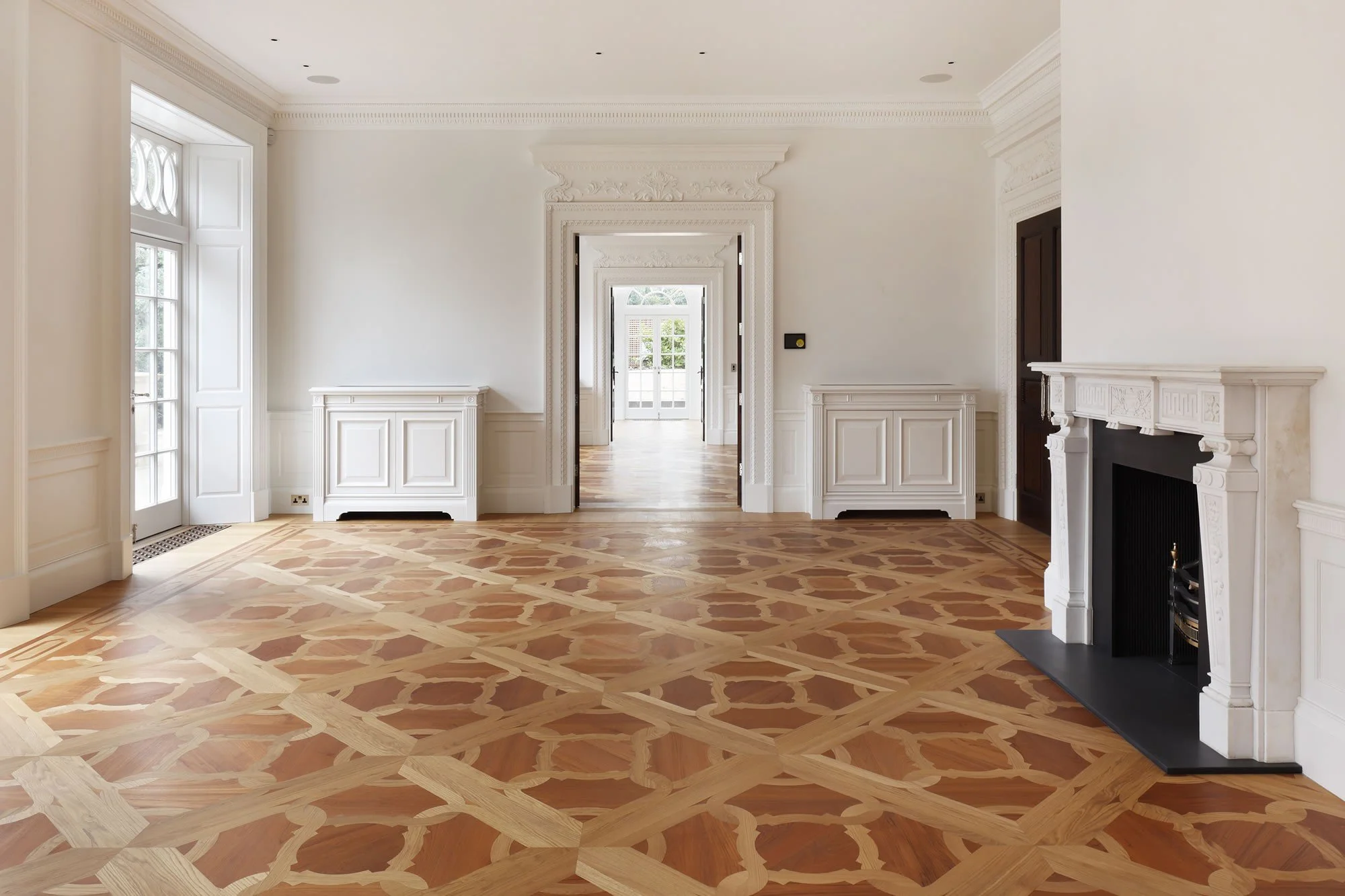 Sala de estar elegante con paredes y molduras blancas, chimenea de mármol y pisos de madera con patrón entrelazado, puertas de vidrio favorecen la entrada de luz natural.