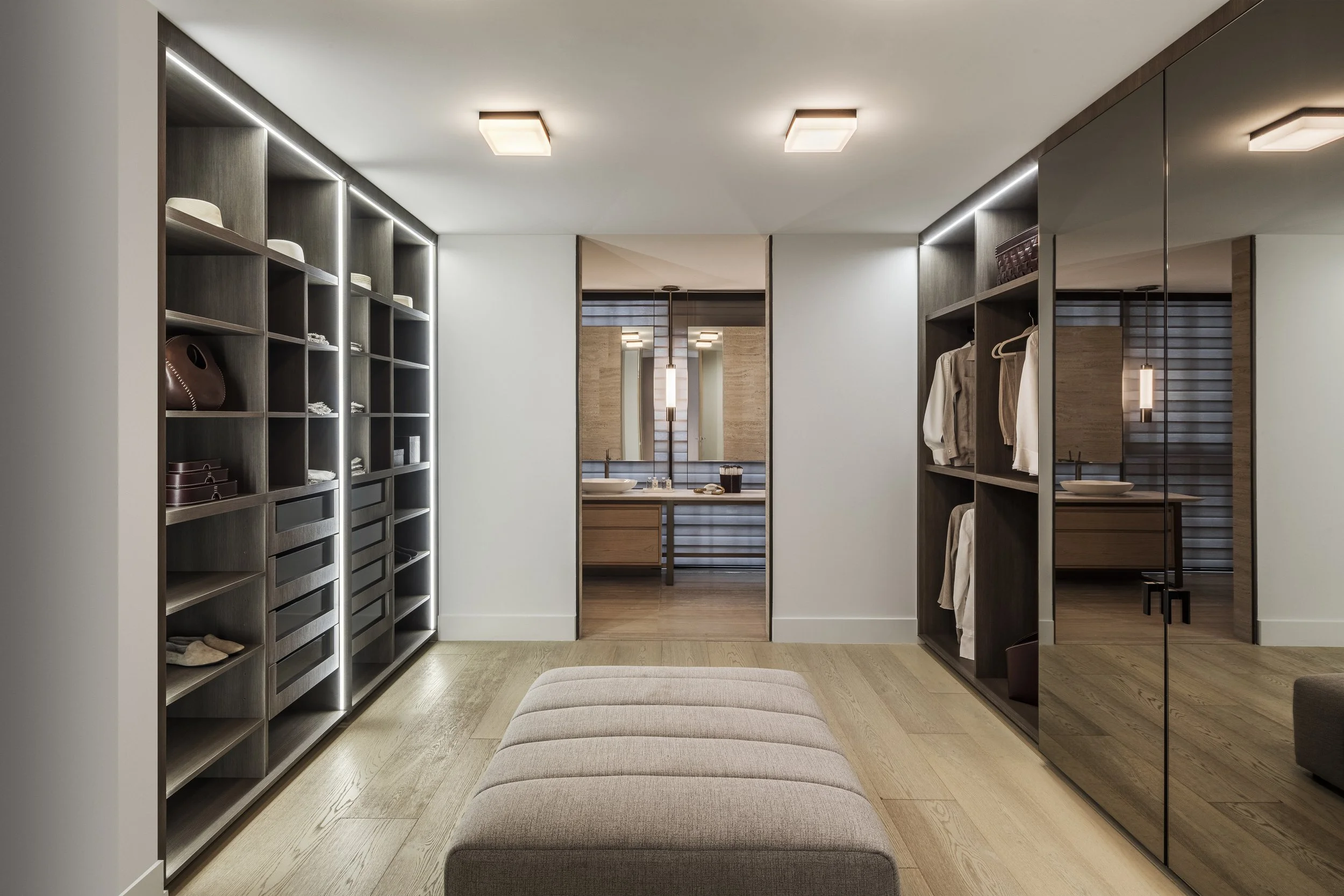 suelos de madera en vestidor moderno con armarios de madera oscura y espejos, un banco acolchado en el centro y un baño en el fondo con lavabos dobles y espejo largo.