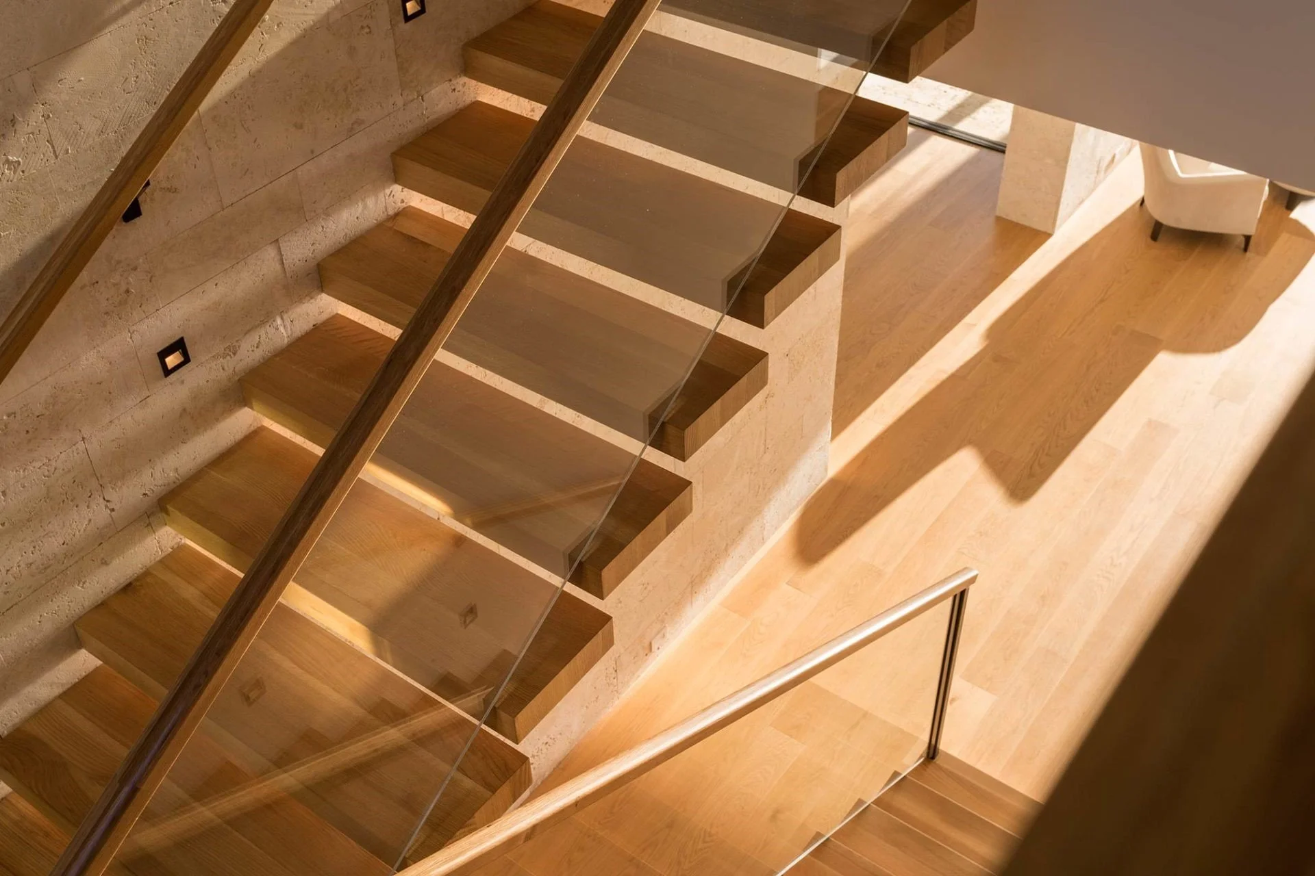 Vista de una escalera moderna de madera y cristal, con paredes de piedra y piso de madera clara, en un interior bien iluminado.