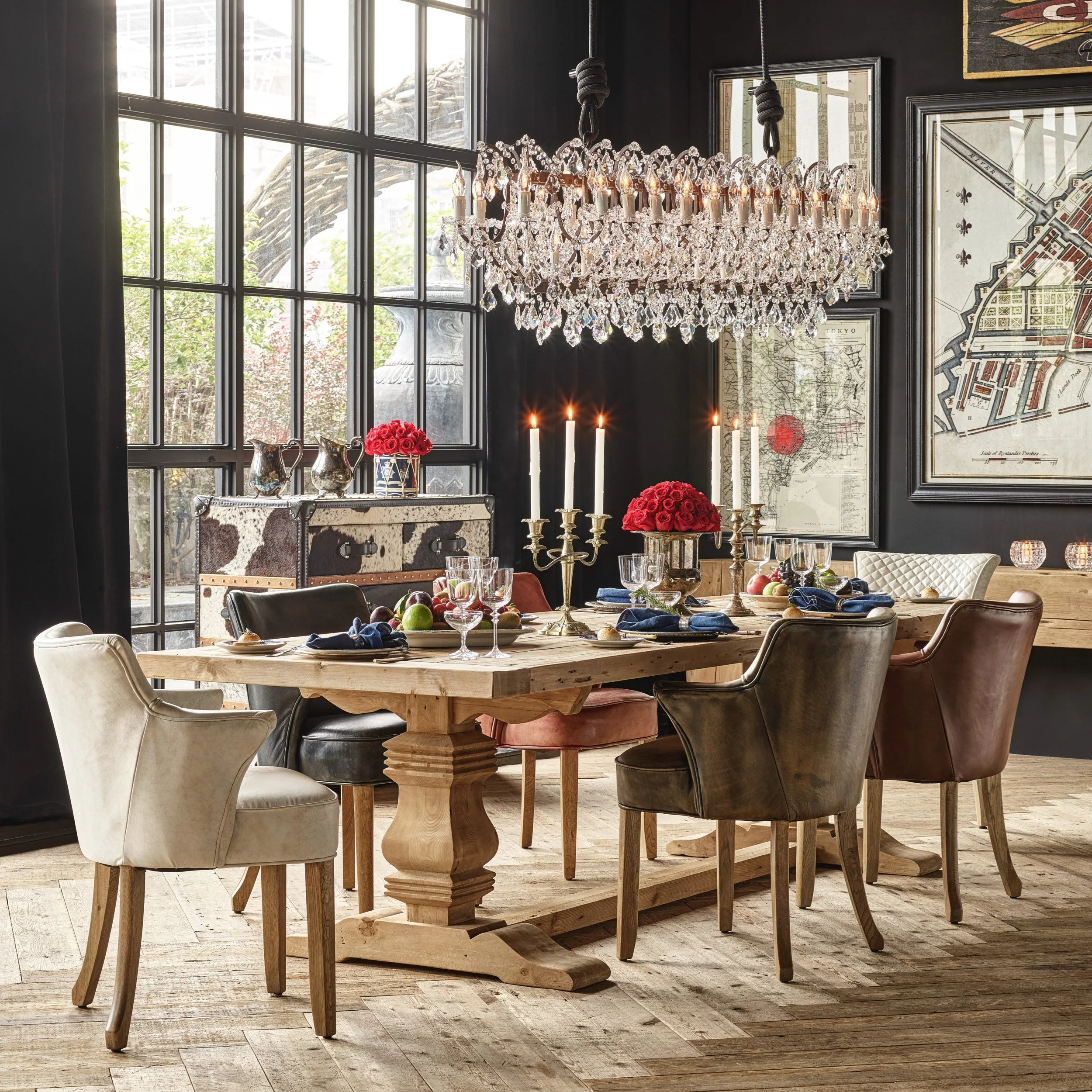 Comedor elegante con mesa de madera rodeada de sillas de diferentes estilos, candelabros con velas encendidas, flores rojas, cristalería y una lámpara de araña de cristales, con paredes negras y ventanas grandes que dejan entrar luz natural.