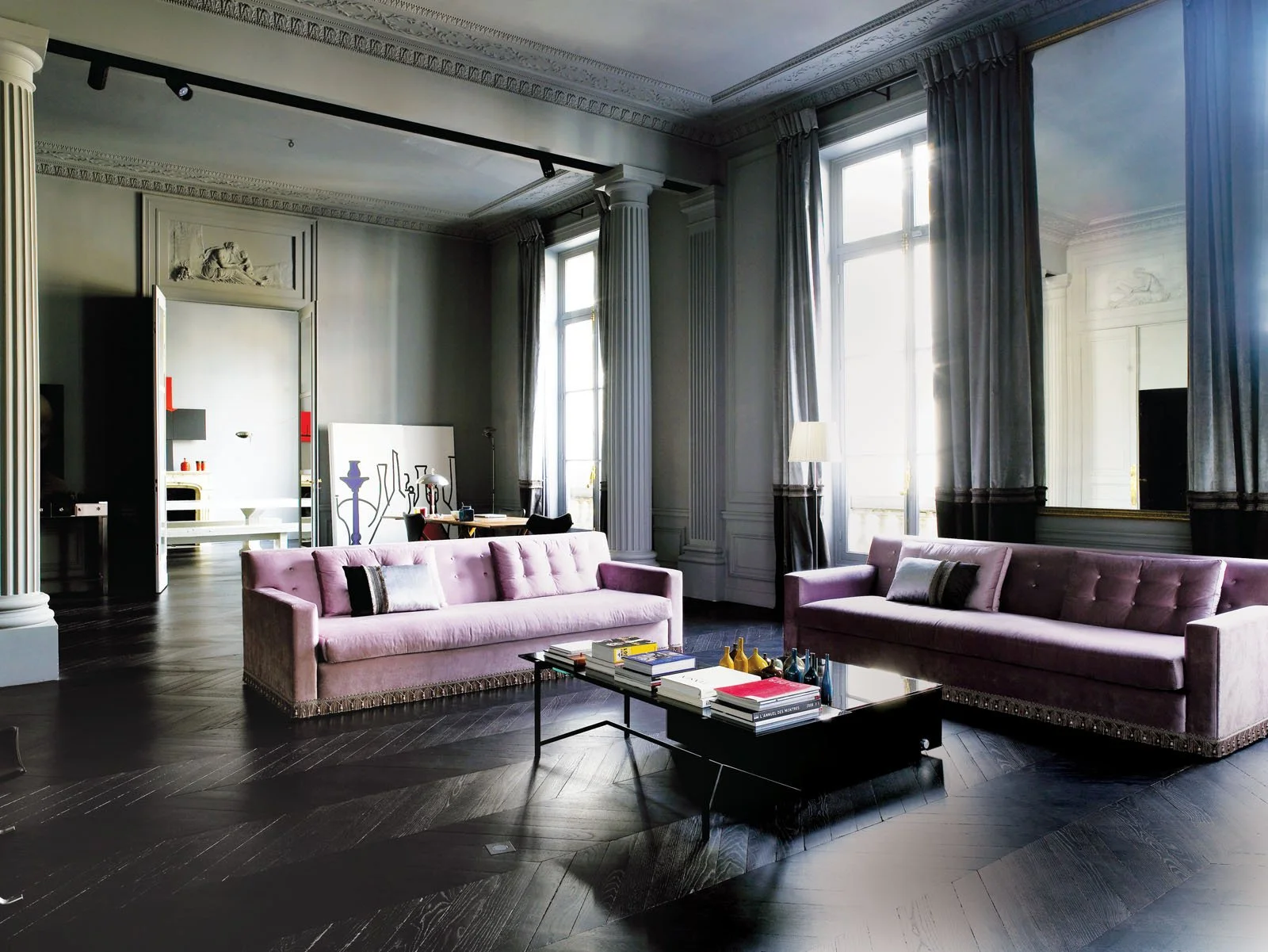 Sala de estar con sillones rosas, mesa de café con libros y objetos decorativos, grandes ventanas con cortinas grises y paredes decoradas con molduras.