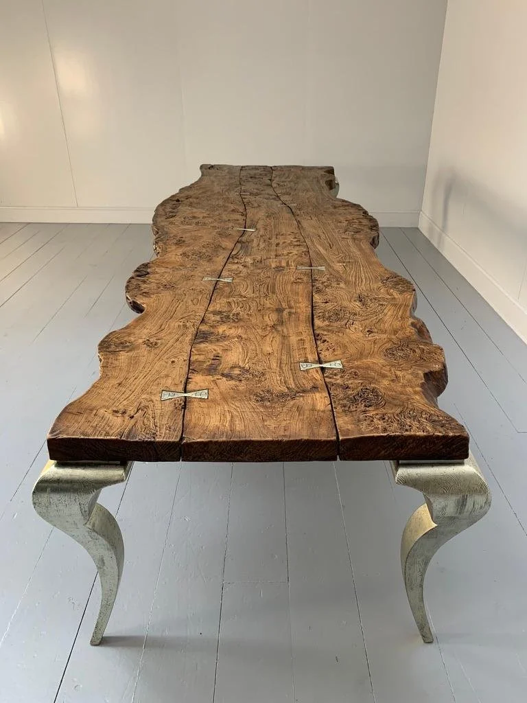 Mesa rústica de madera con patas curvas de color plateado, en una habitación con paredes blancas y piso claro.