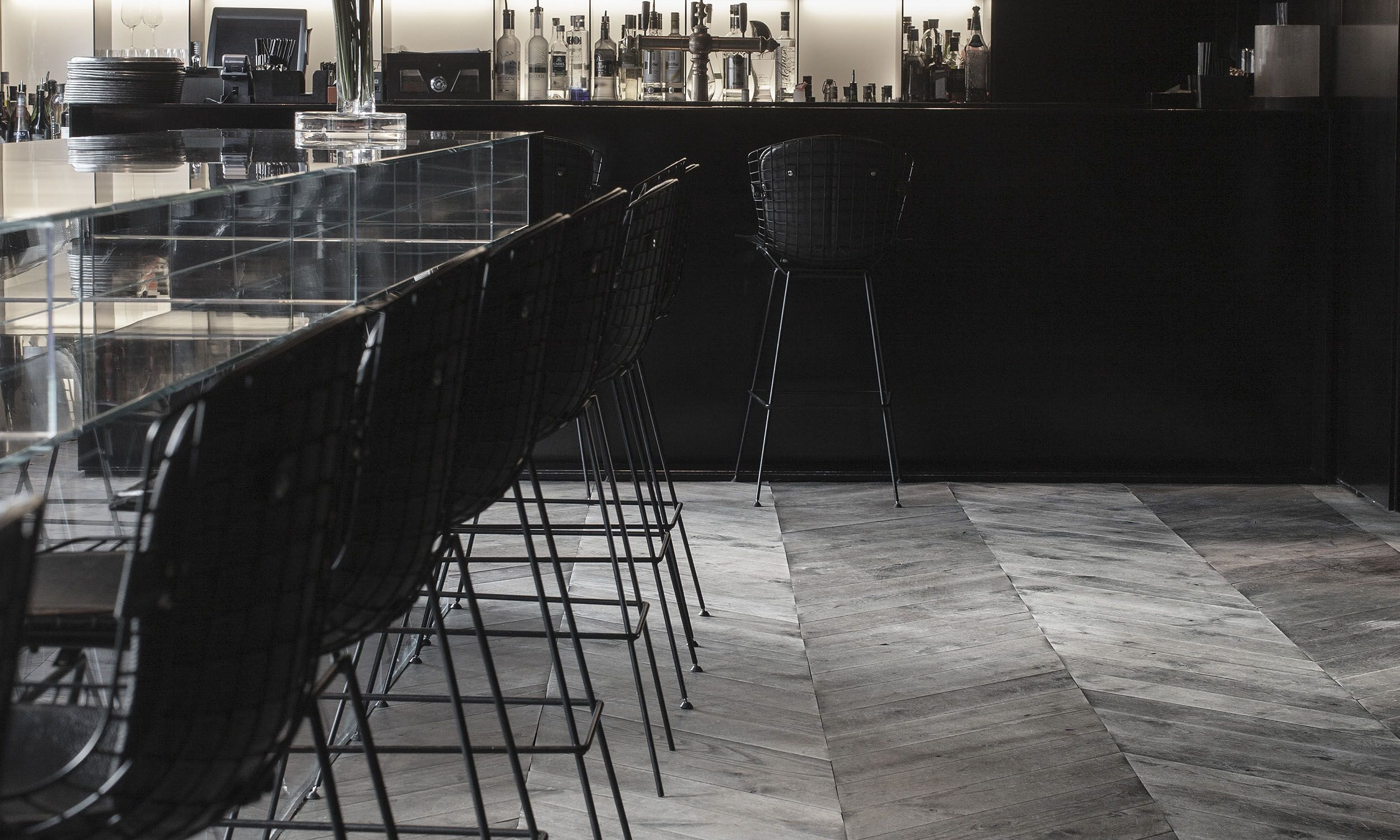 Bar con sillas de malla negras alrededor, estantería con botellas y vasos, suelo de madera, ambiente moderno y minimalista.