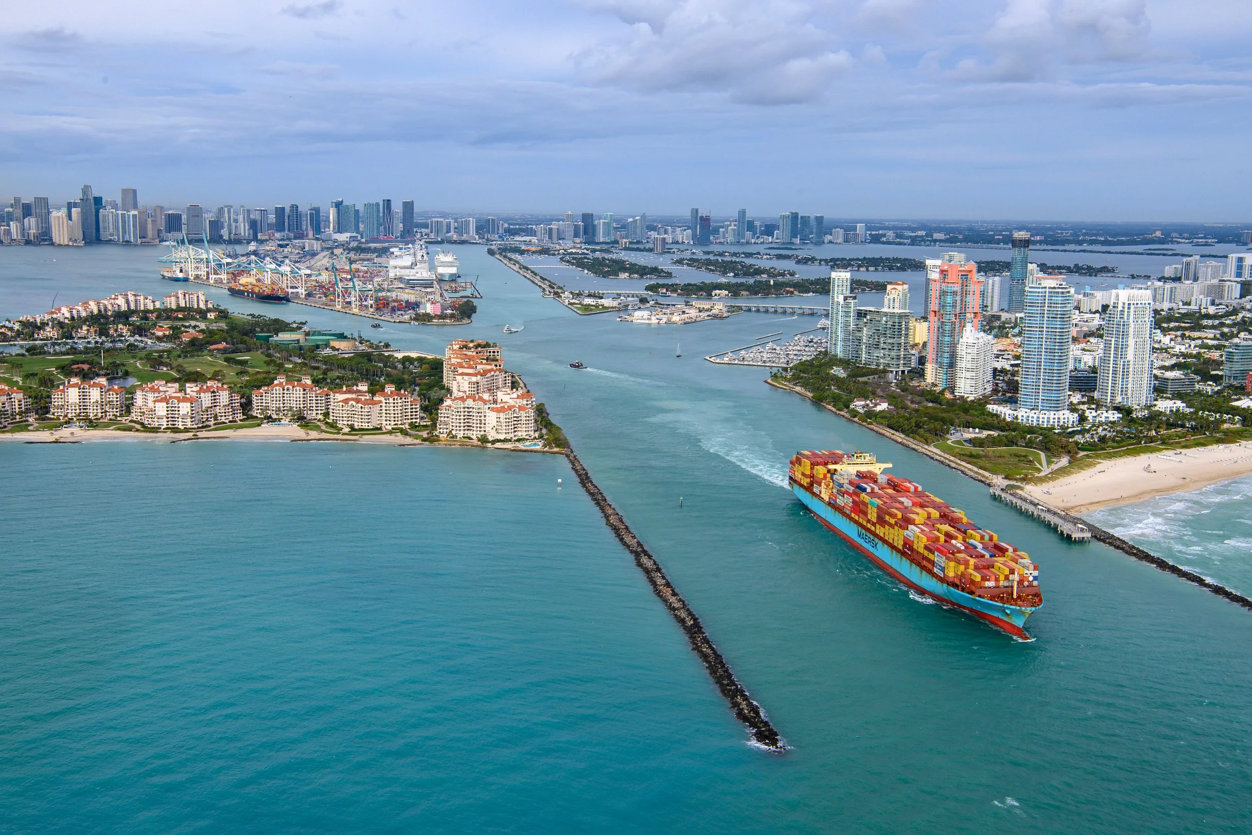 Aerial view of a busy port in Miami with a container ship sailing through a canal, high-rise buildings, a beach, and the city skyline in the background.