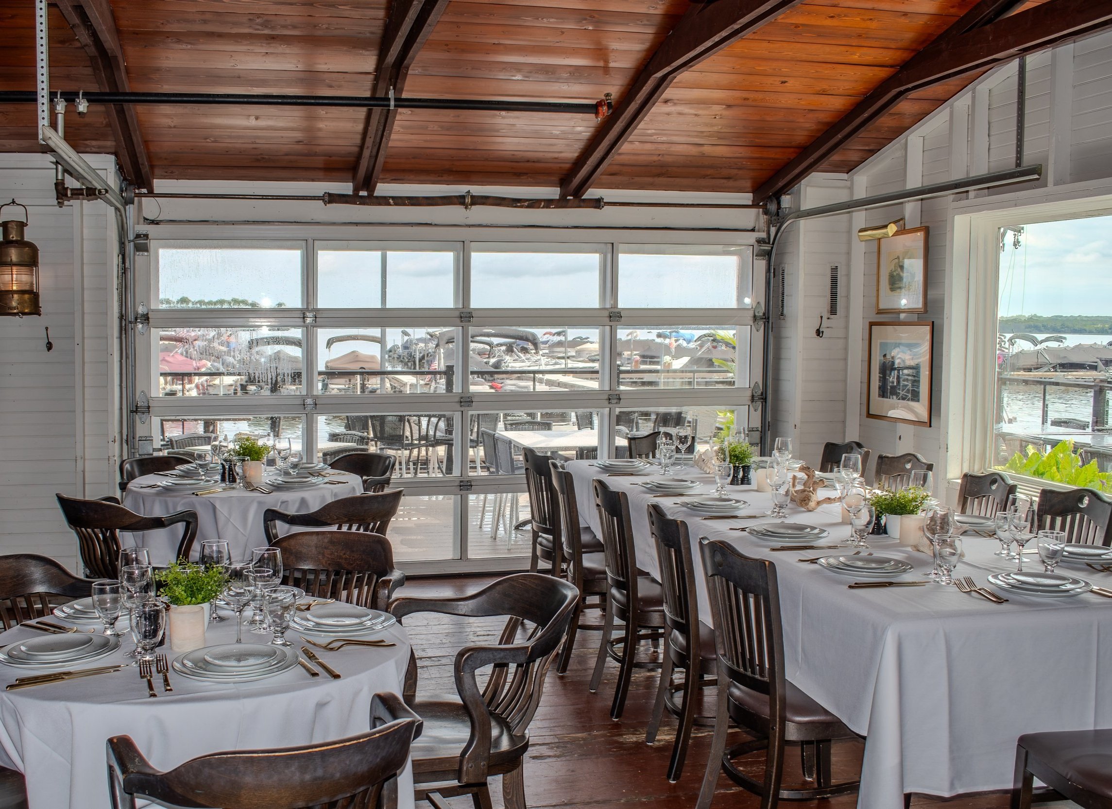 Indoor private dining area with round tables draped in white tablecloths, set with plates, glasses, silverware, and small potted plants, with a large garage-style window revealing boats and a marina outside, and a wooden ceiling with exposed beams.