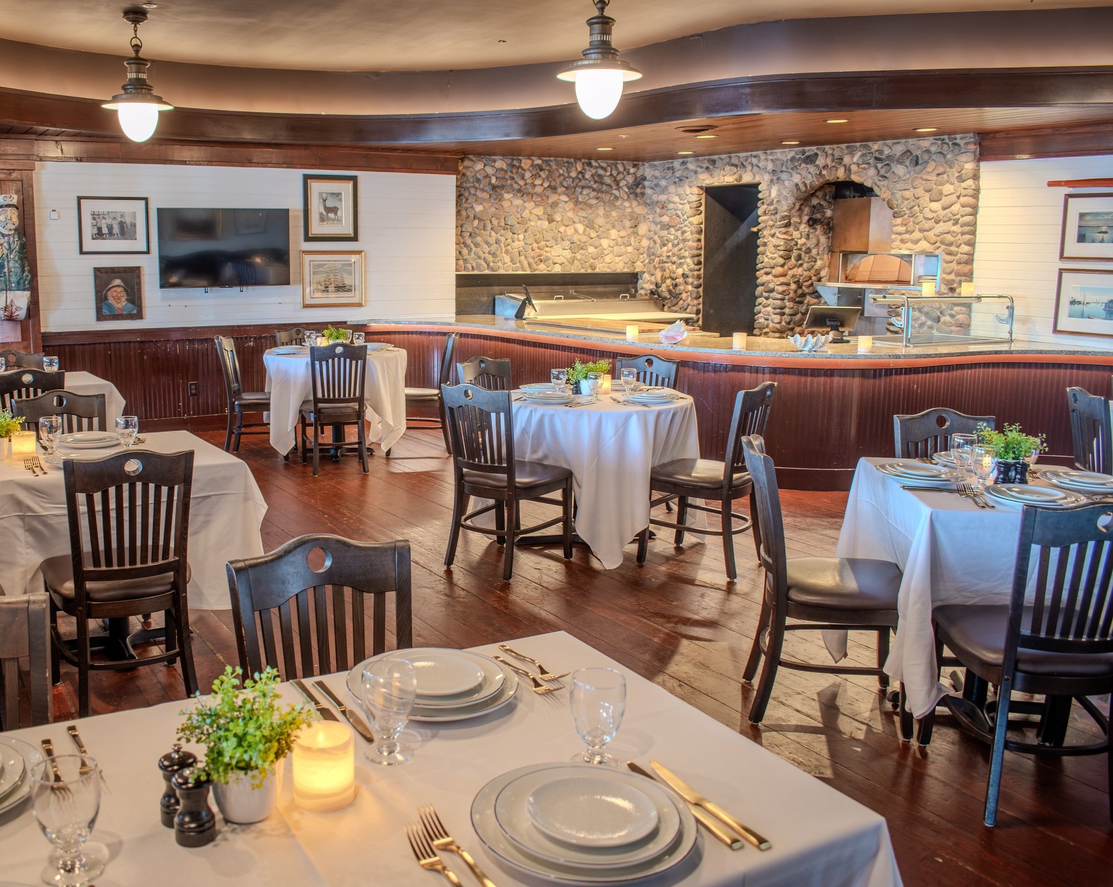 Elegant restaurant interior with wooden accents, stone wall, and tables set with white tablecloths, plates, glasses, and cutlery, decorated with candles and green plants for private dining.