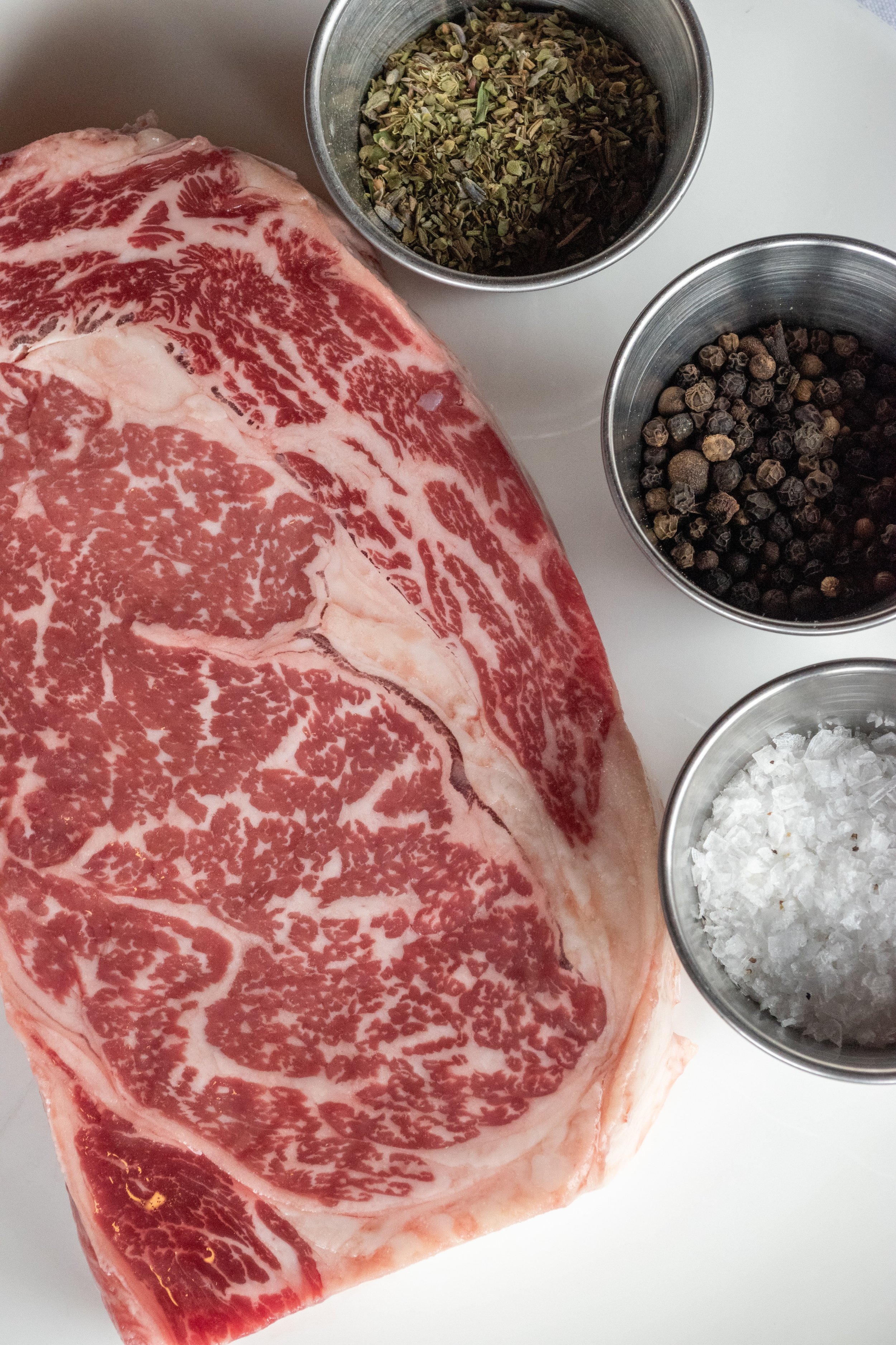 A close-up of a large, marbled piece of raw Eden Valley Farms wagyu beef steak alongside three small metal bowls containing dried herbs, black peppercorns, and coarse sea salt on a white surface.