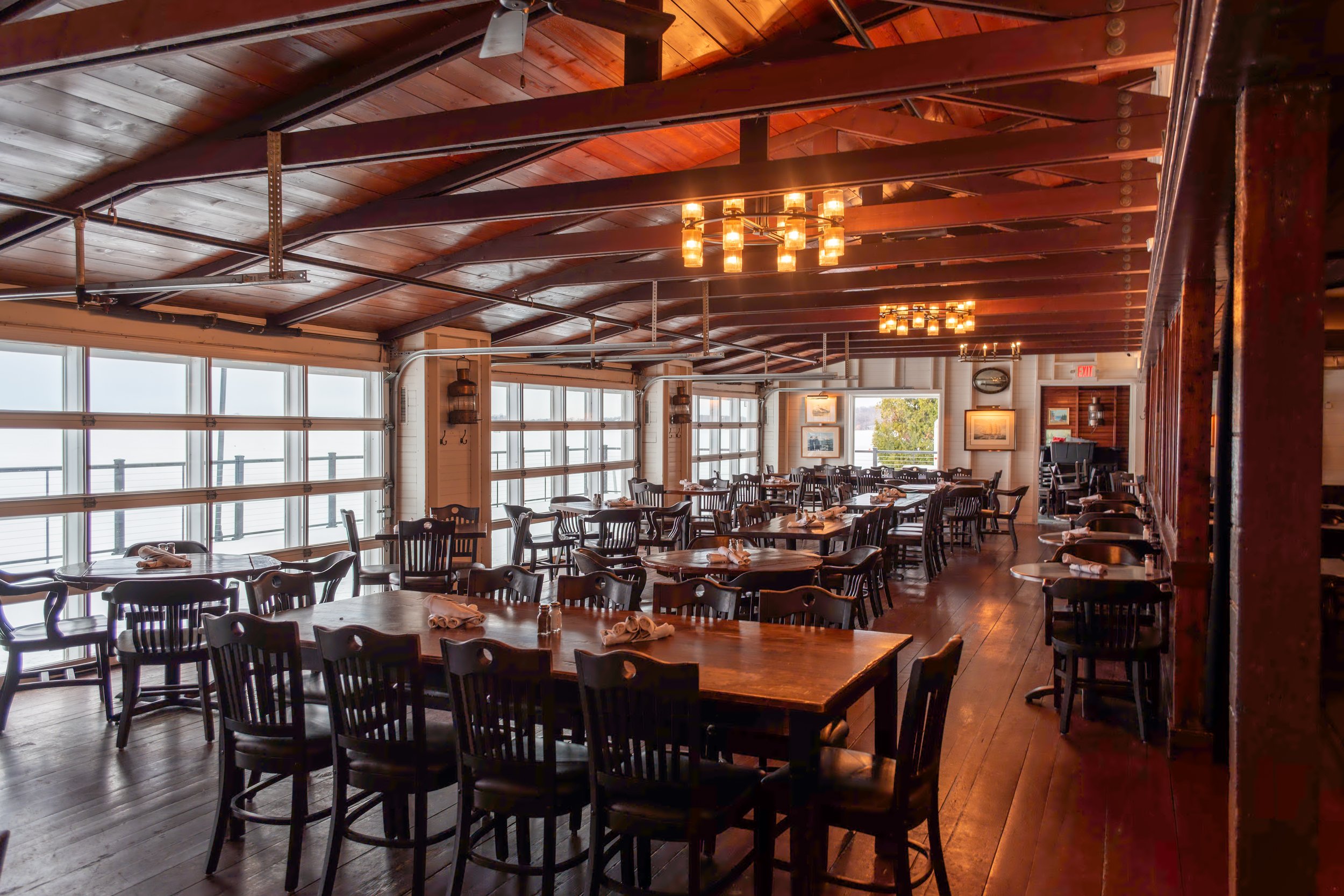 Interior of a rustic restaurant with wooden ceiling and beams, large windows, hanging lights, and wooden tables and chairs set for private dining.