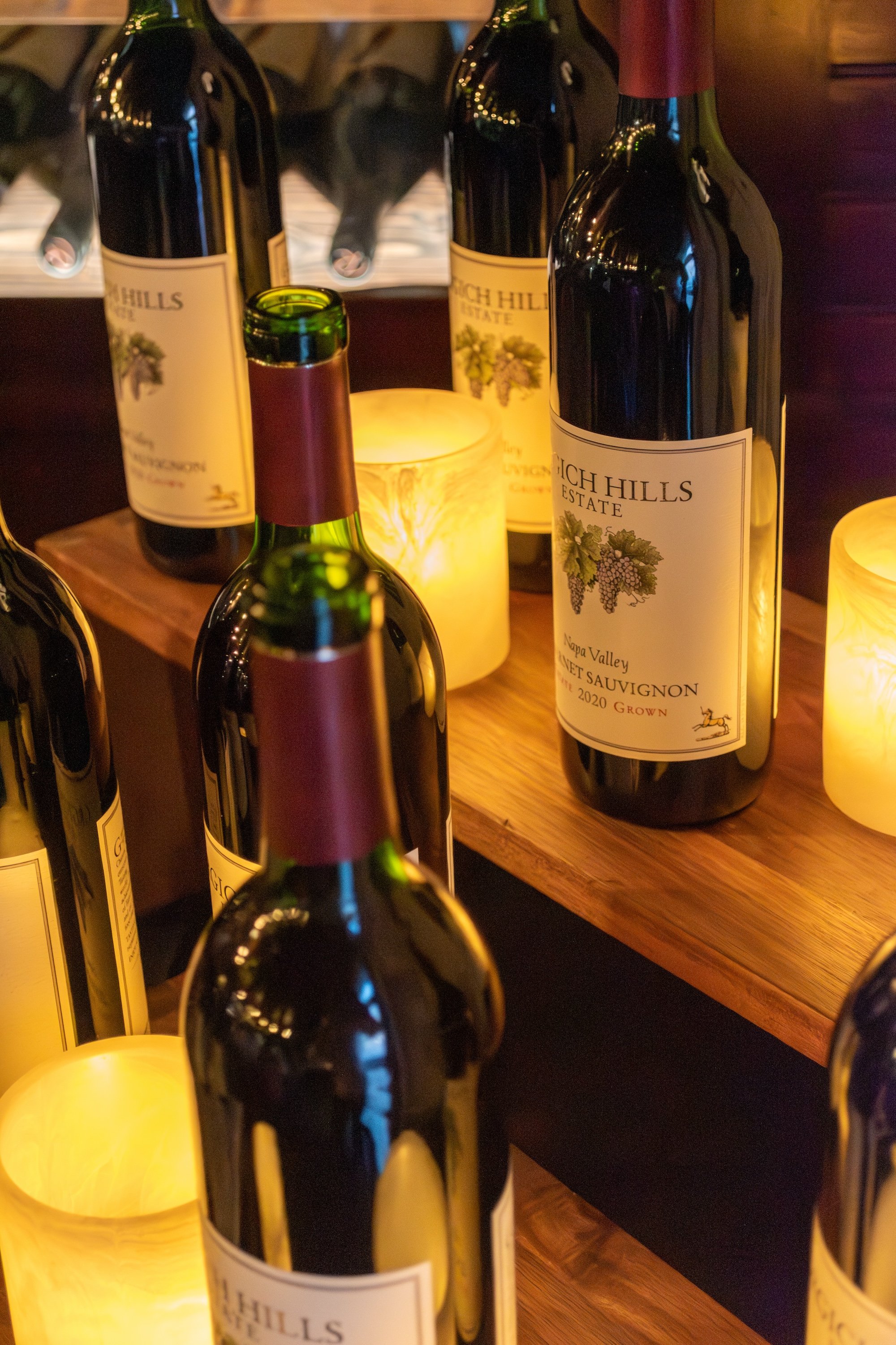 Several bottles of wine on a wooden shelf with small glowing candle lights around them.
