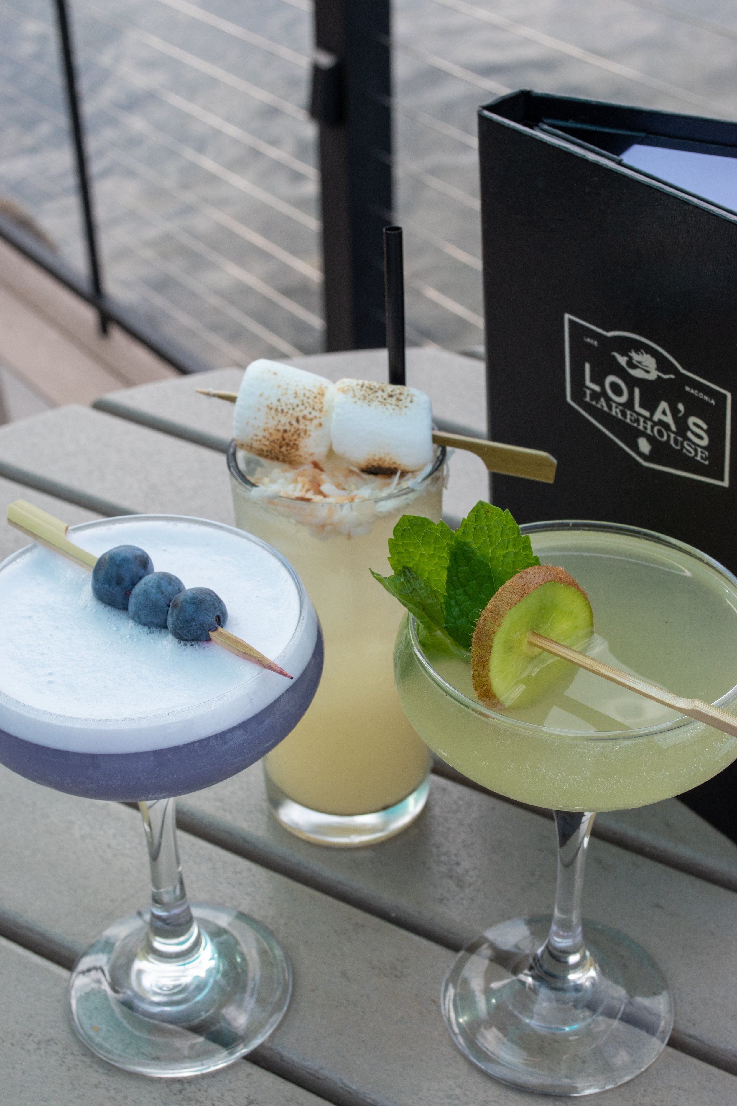 Three cocktails are placed on a table, two are with stemmed glasses and one is with a short glass, decorated with fruits and marshmallows. A shopping bag from Lola's Lakehouse is in the background.
