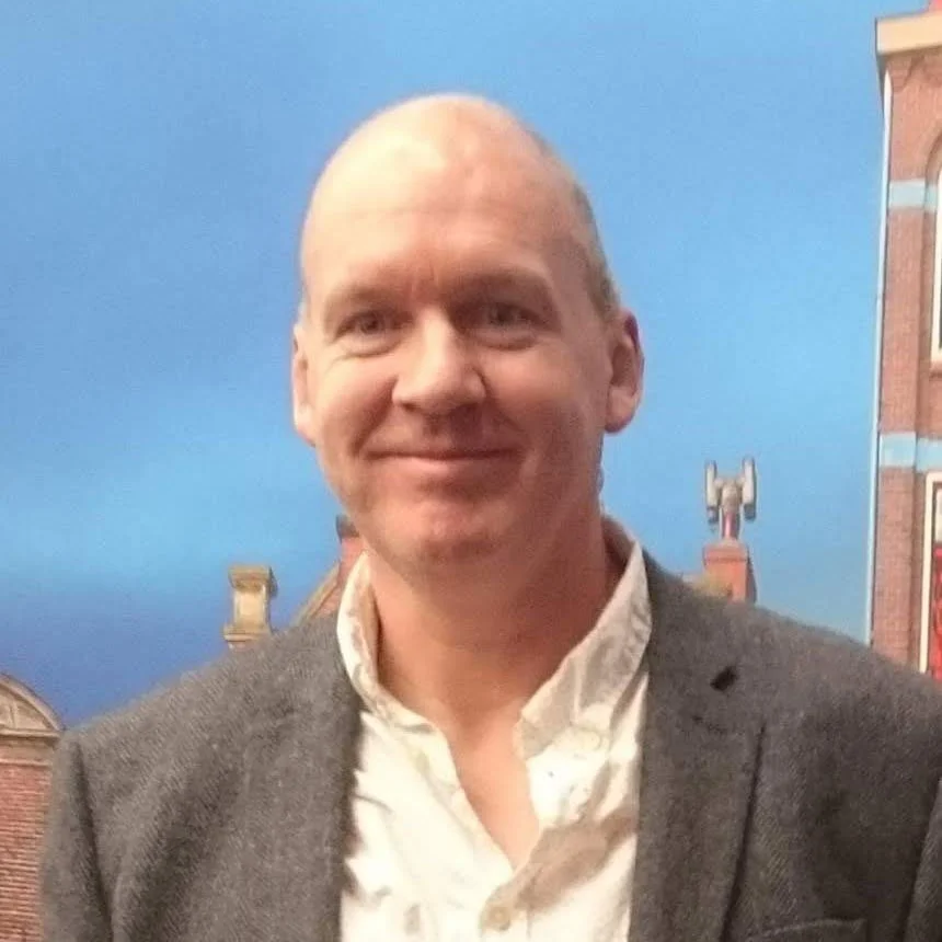 A smiling man with a bald head wearing a gray blazer and white shirt, standing in front of a painted backdrop of a blue sky and brick buildings.