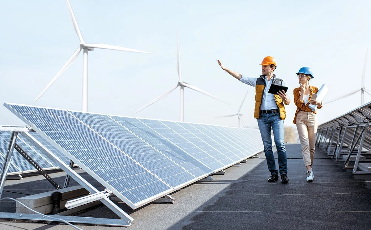 Dos ingenieros en casco y gafas revisando paneles solares y aerogeneradores en un parque eólico