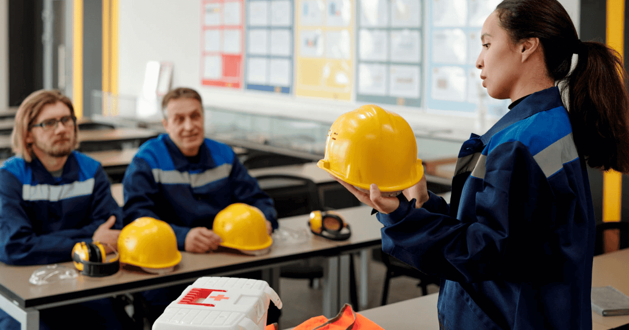 Mujer en uniforme de seguridad mostrando un casco de construcción a un grupo de personas en un aula de formación de seguridad laboral.