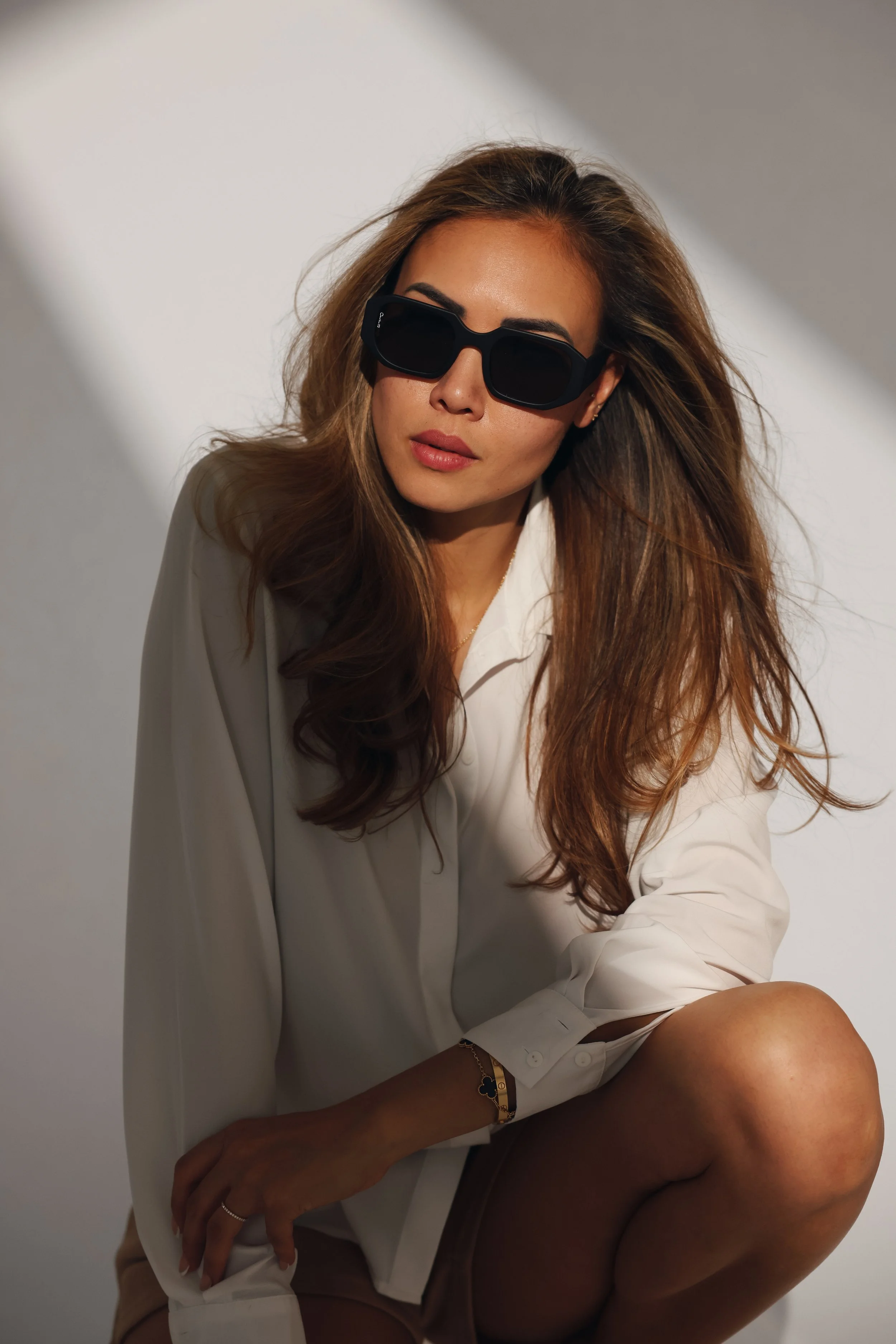 A woman with long wavy hair, wearing large black sunglasses and a white shirt, poses against a neutral background.