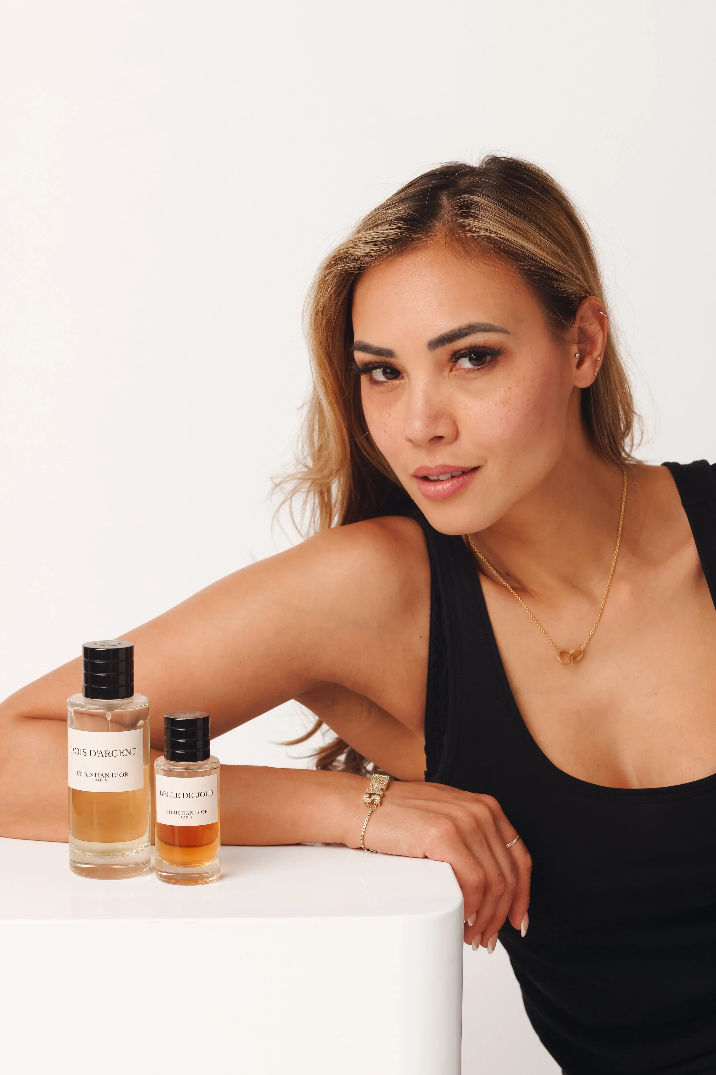 A woman with light brown hair, wearing a black top and gold jewelry, is posing next to two bottles of Christian Dior perfume on a white surface against a plain off-white background.