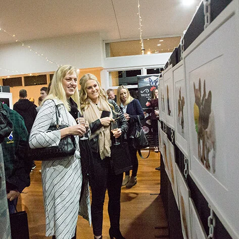 Two women smiling and looking at artwork displayed on a wall at an indoor event.