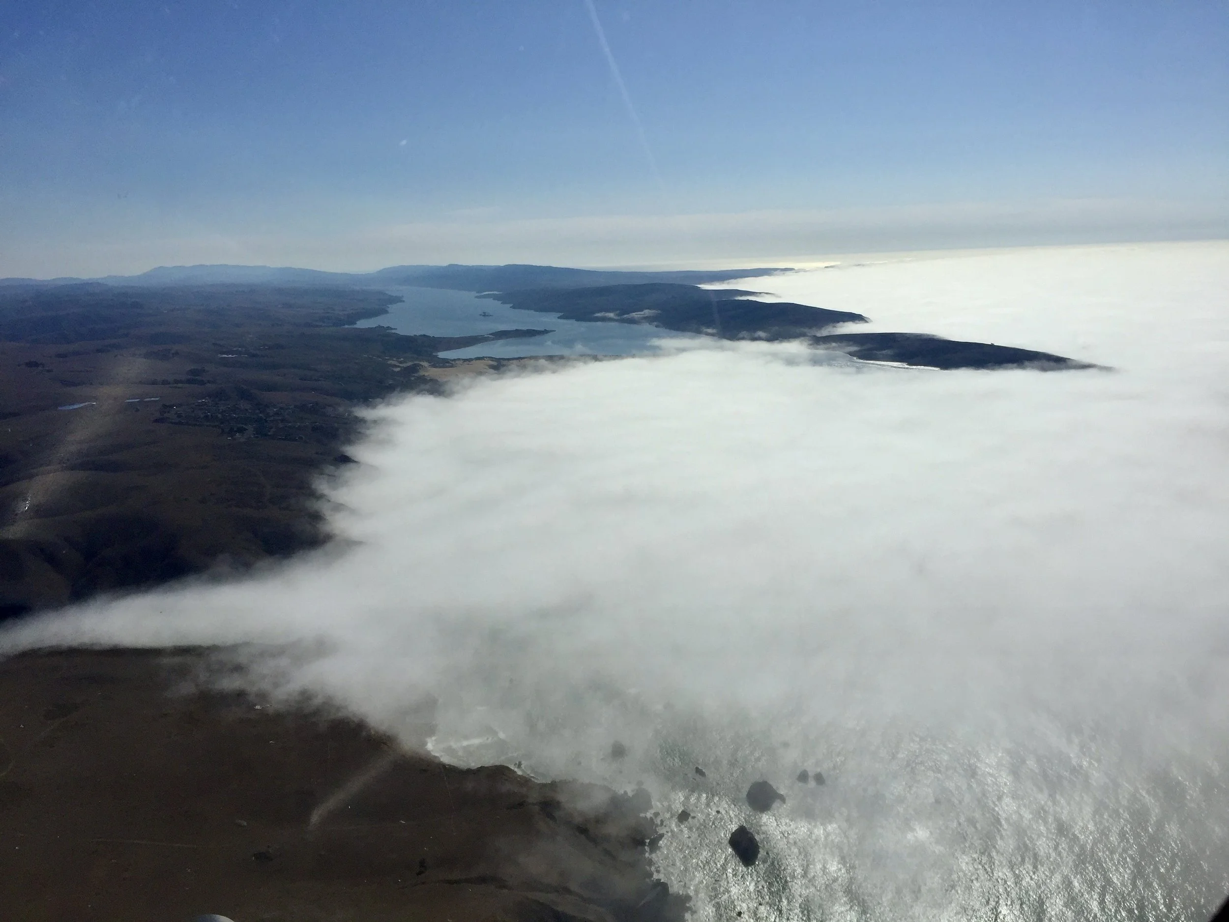 Aerial view of a landscape with a large body of water and shoreline partially obscured by low-hanging clouds under a clear blue sky.
