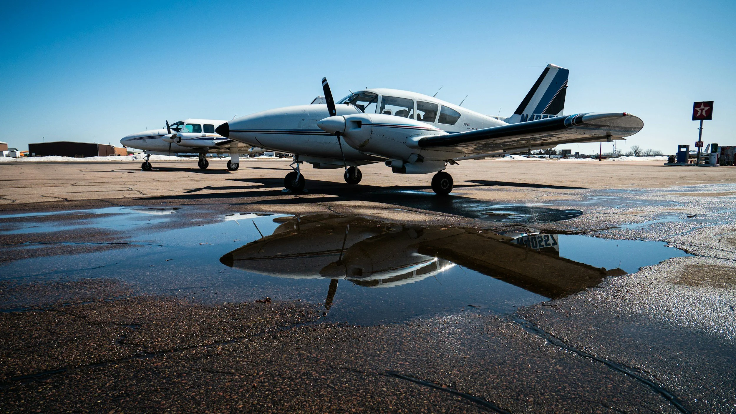Small white airplane parked on an airport tarmac, with its reflection visible in a puddle of water in the foreground. Additional airplanes and a gas station are visible in the background.