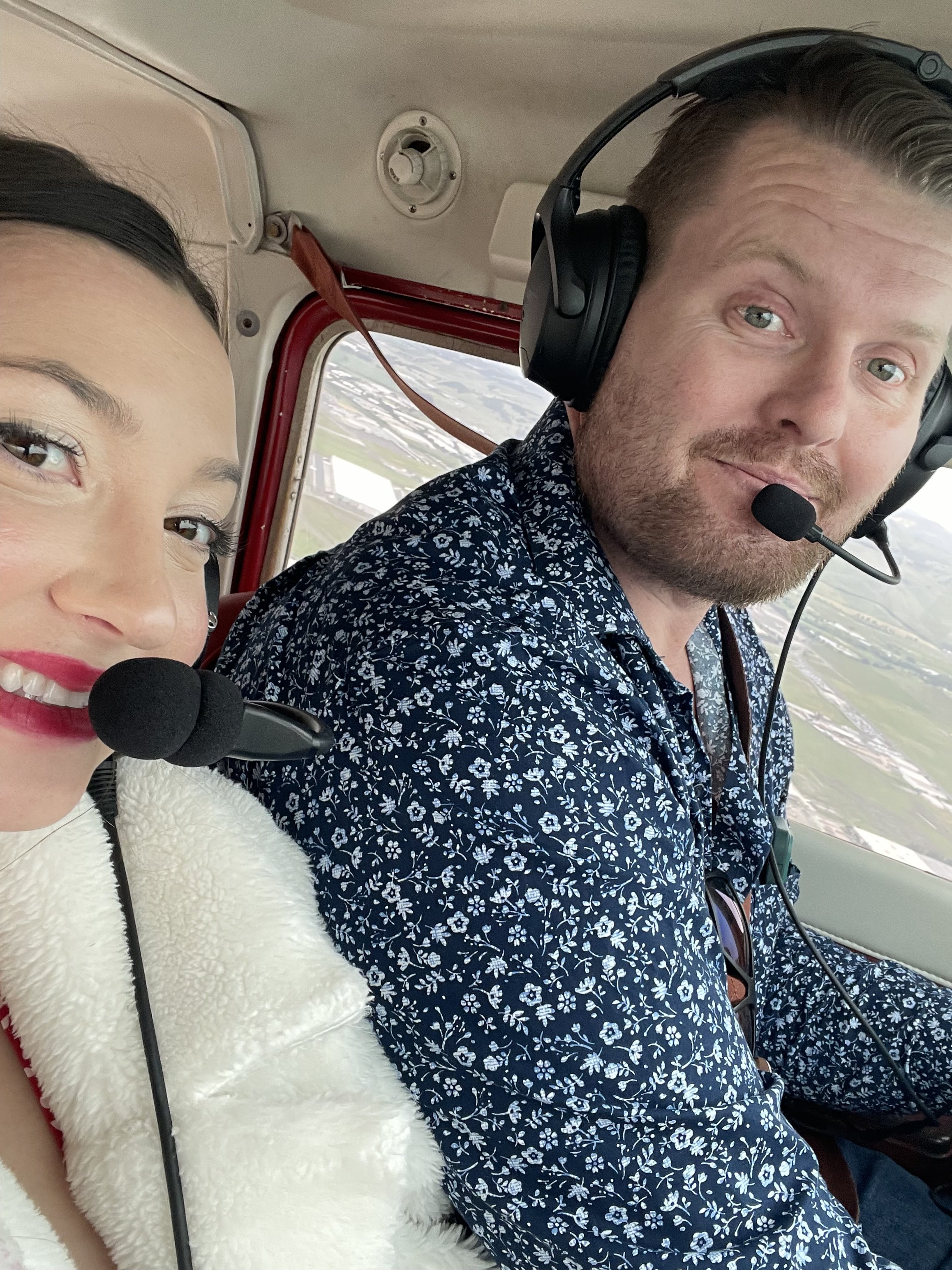 Two people smiling in the cockpit of a small aircraft, wearing headsets, with a view of the landscape below through the window.