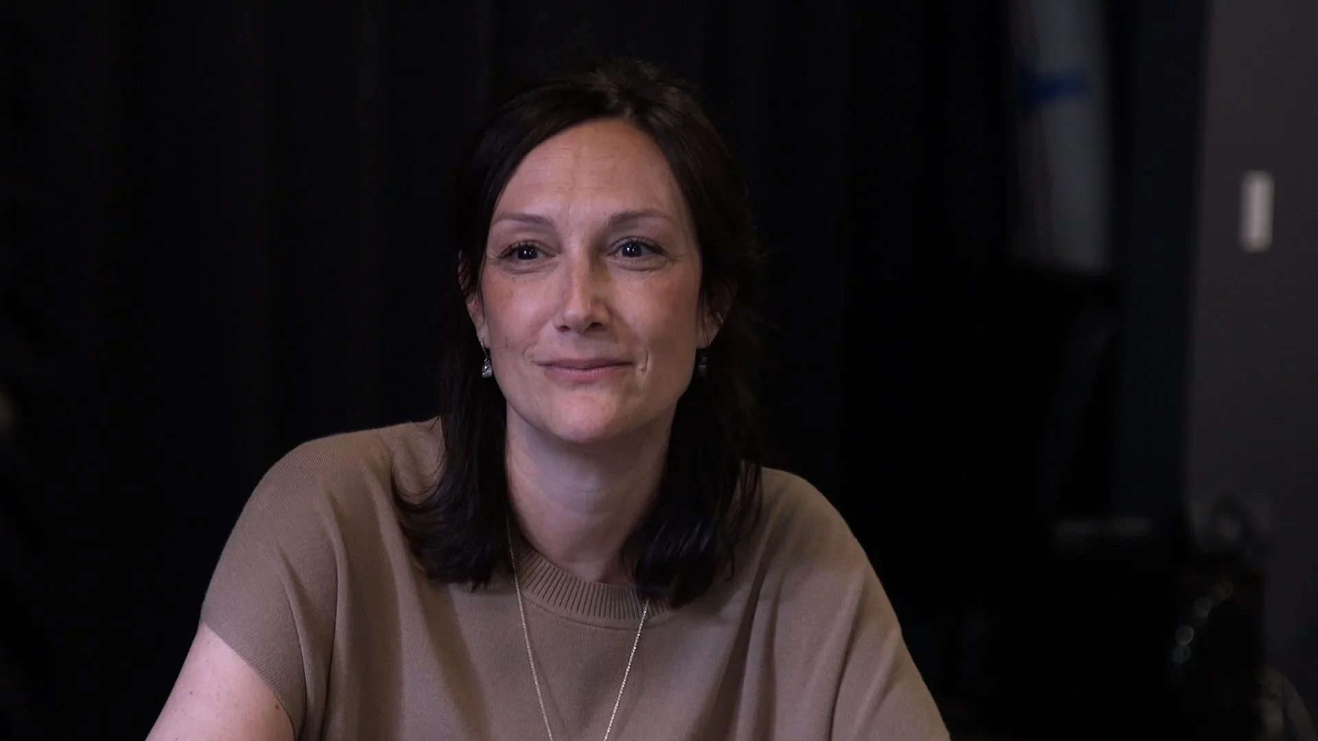A woman with dark hair and earrings sitting at a table in front of a black background.