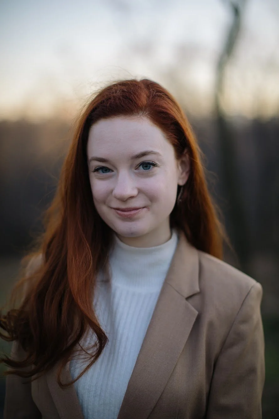 Portrait of a woman with long red hair wearing a beige blazer and white turtleneck, outdoors during sunset.