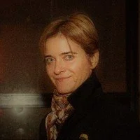 A woman with short brown hair wearing a black top and a patterned scarf, smiling in a dimly lit setting.