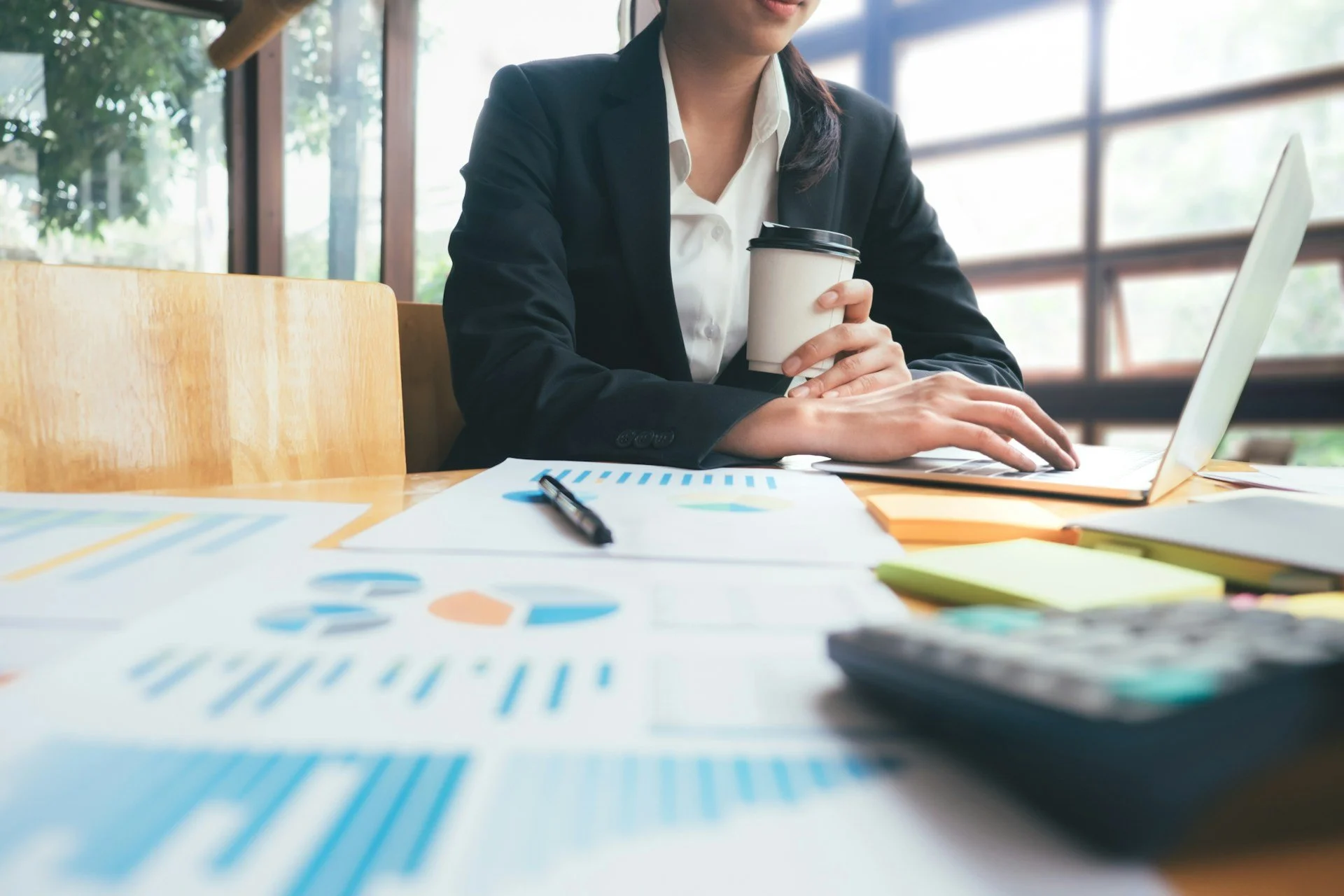 A woman in a business suit sitting at a table with a laptop, holding a coffee cup, with financial graphs, charts, a pen, sticky notes, and a calculator on the table.