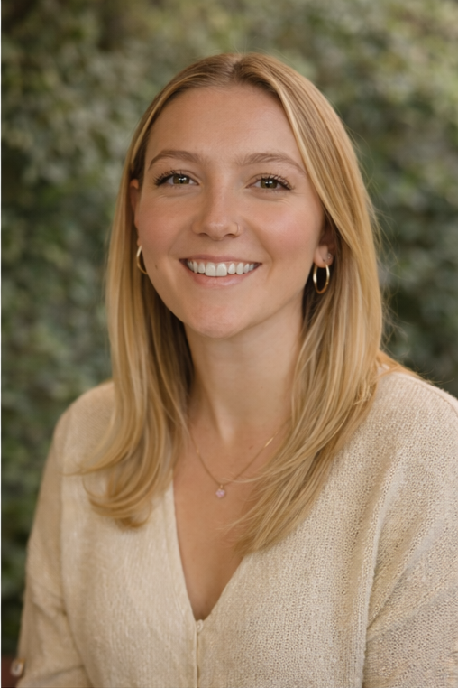 Portrait of a young woman with blonde hair, smiling, wearing hoop earrings, a small necklace, and a light-colored sweater, with a blurred outdoor background.