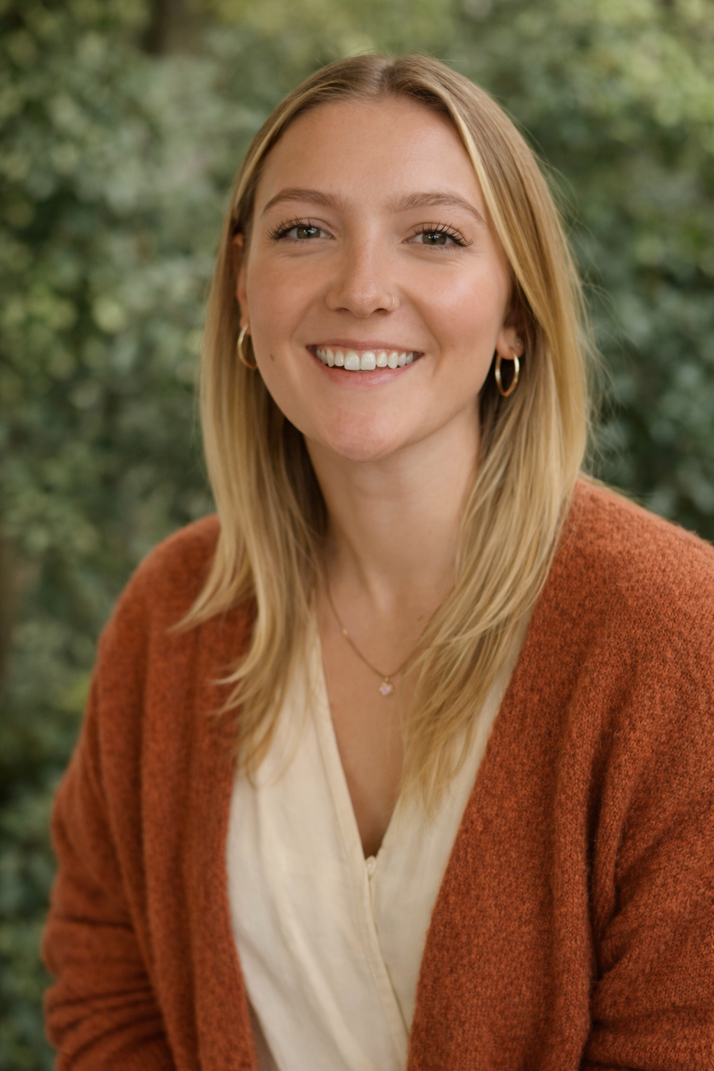 A young woman with blonde hair, wearing a rust-colored sweater over a cream blouse, smiling outdoors with green blurred foliage in the background.