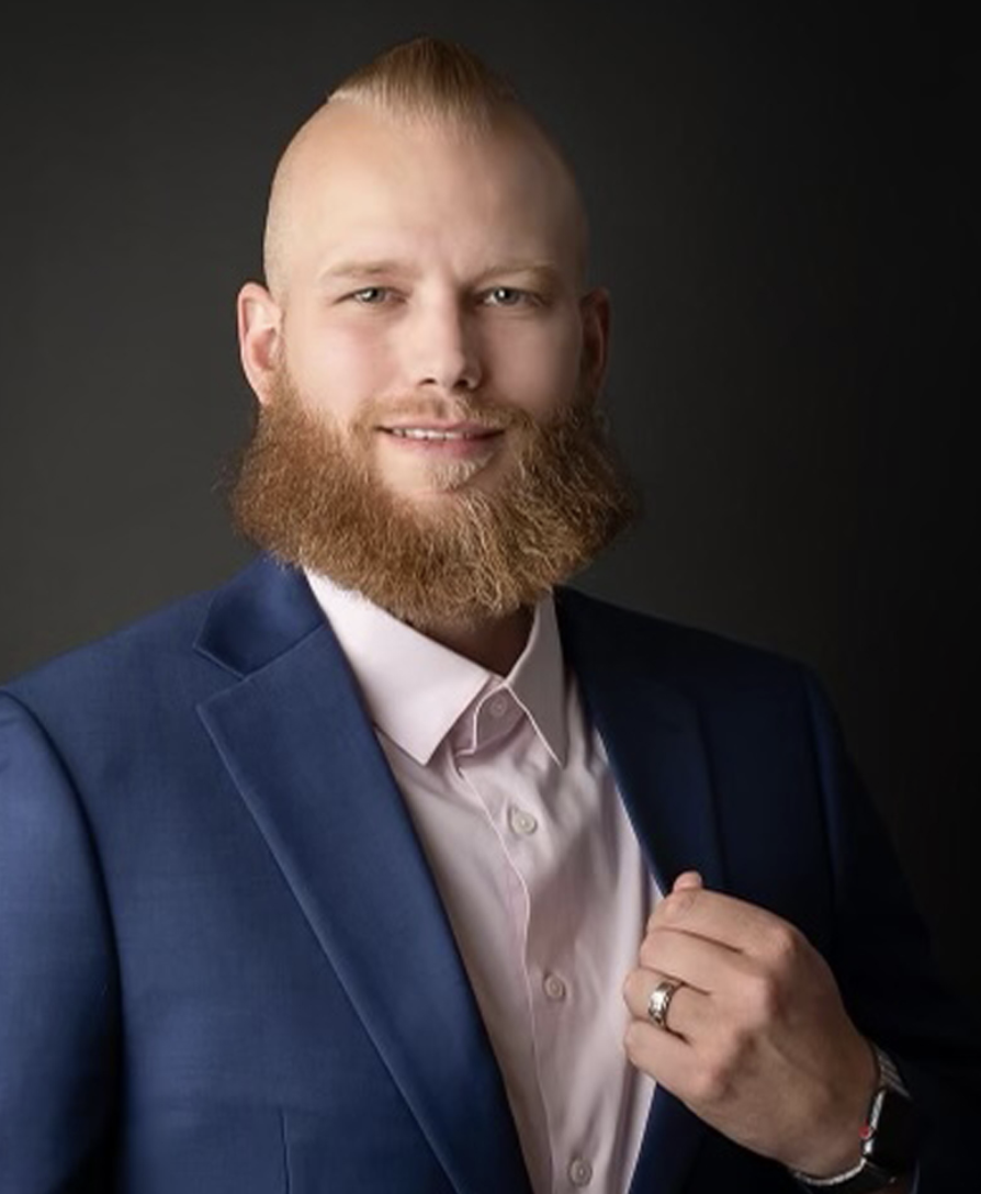 A man with a beard in a suit and pink shirt, posing against a dark background.