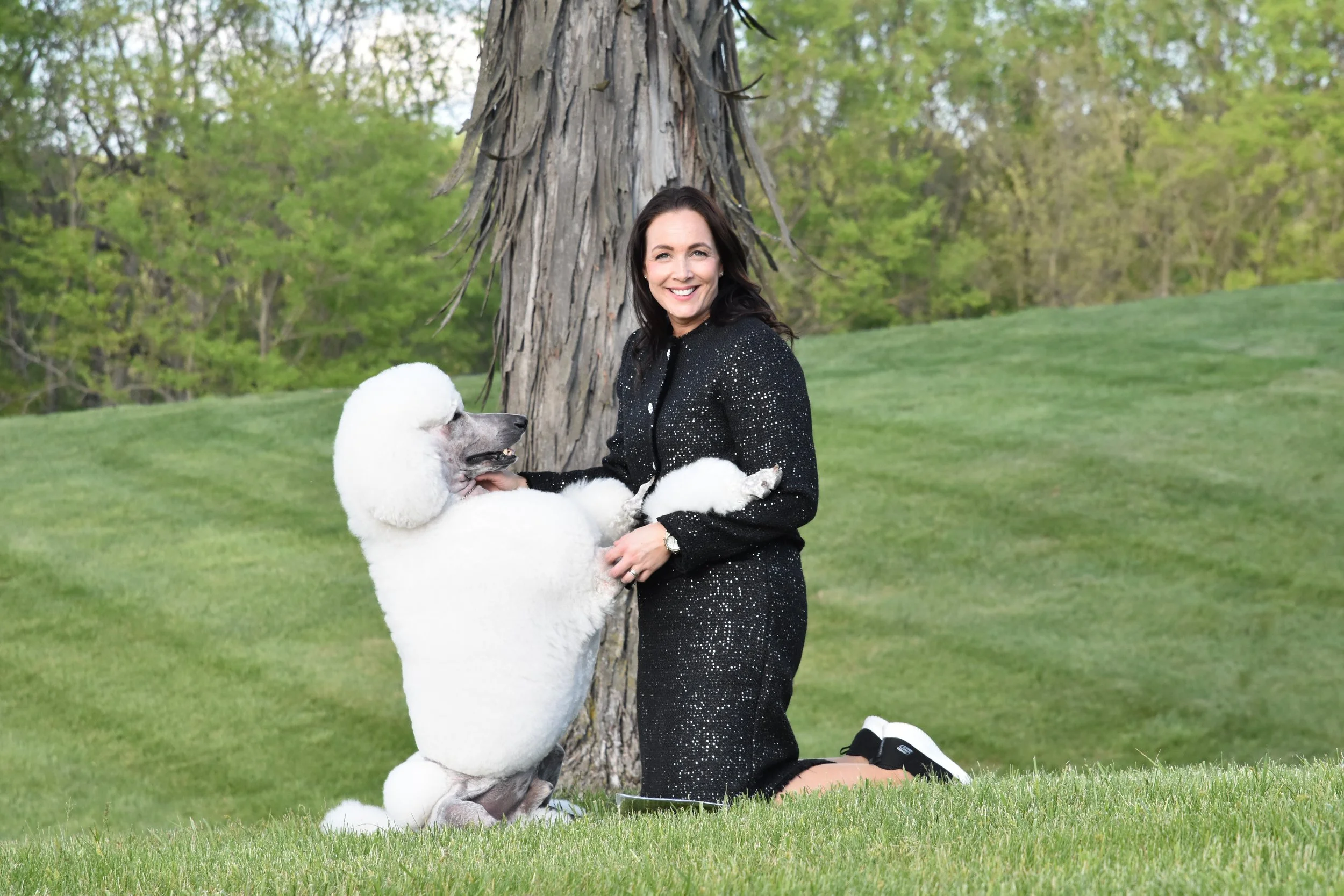 A woman kneeling on the grass next to a large poodle with a fluffy white coat, both outdoors near a tree, smiling at the camera.