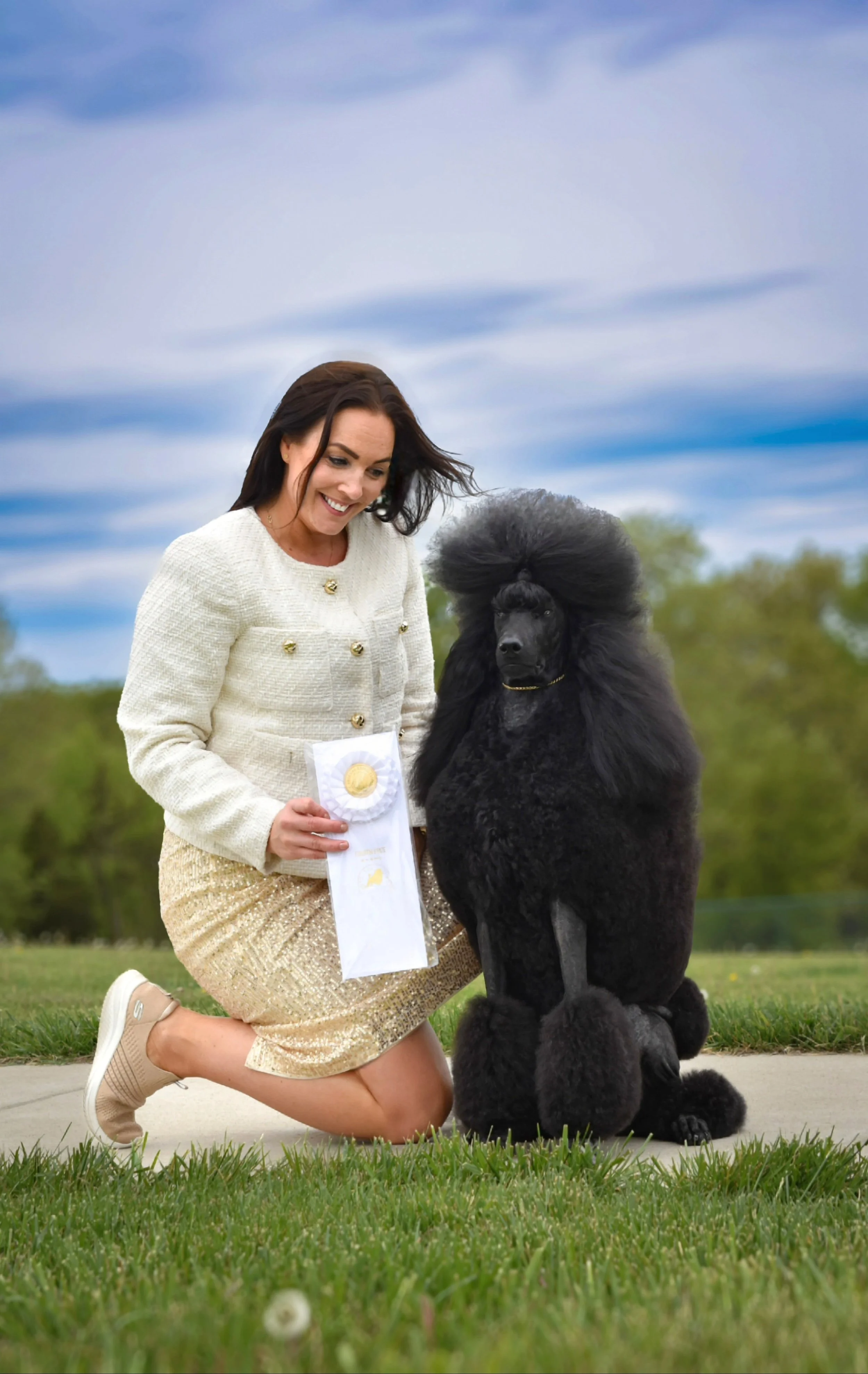 A woman in a white and gold outfit kneels on the grass next to a black poodle with a traditional show haircut, holding a ribbon and smiling at the dog with a blue sky and trees in the background.