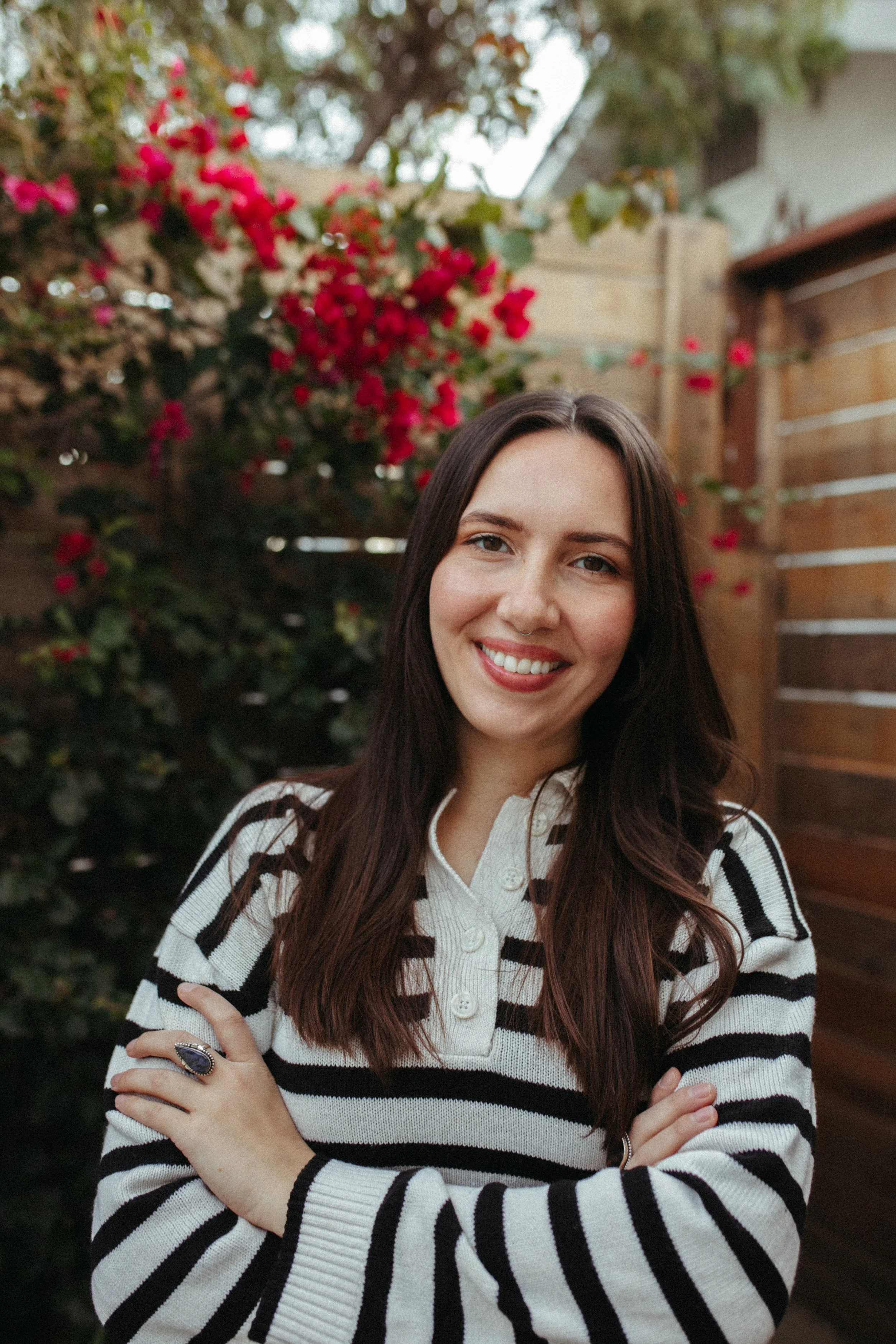 A woman with long dark hair smiling outdoors near a garden with pink flowers and a wooden fence.