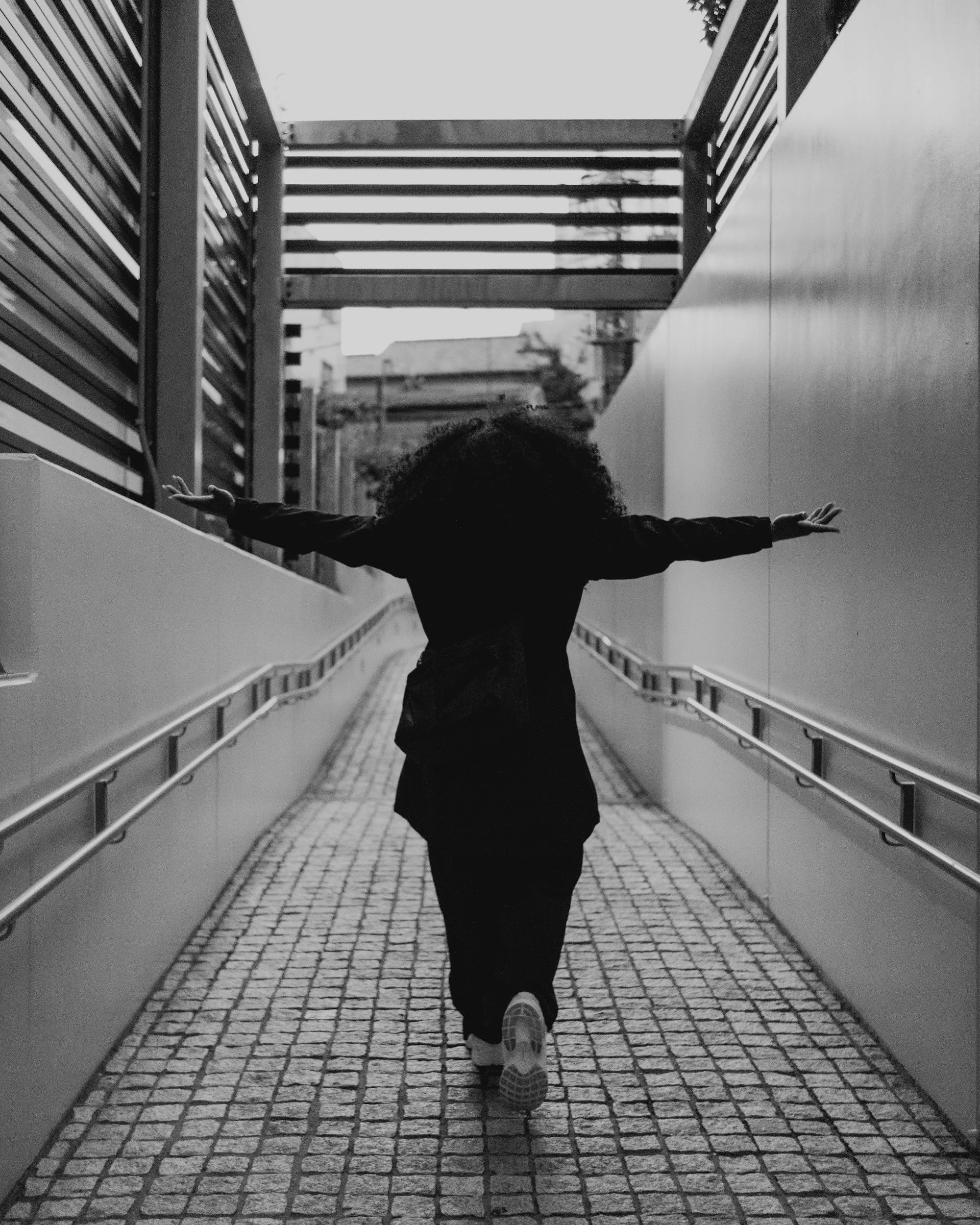 A person with curly hair walking on a cobblestone path with arms outstretched, flanked by metal railings, in an urban setting.