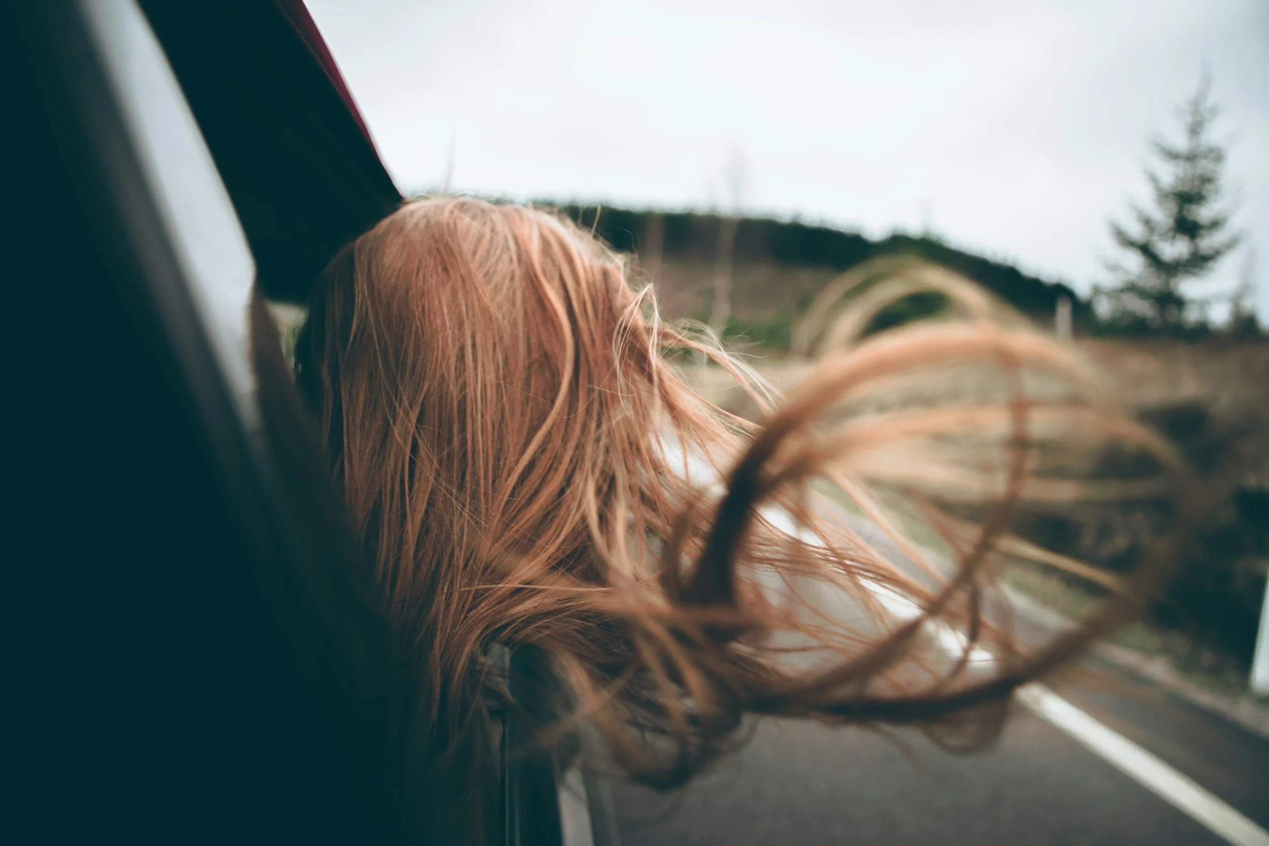 A woman with long, red hair leaning out of a car window, wind blowing her hair, as the car drives along a rural road with trees and overcast sky in the background.