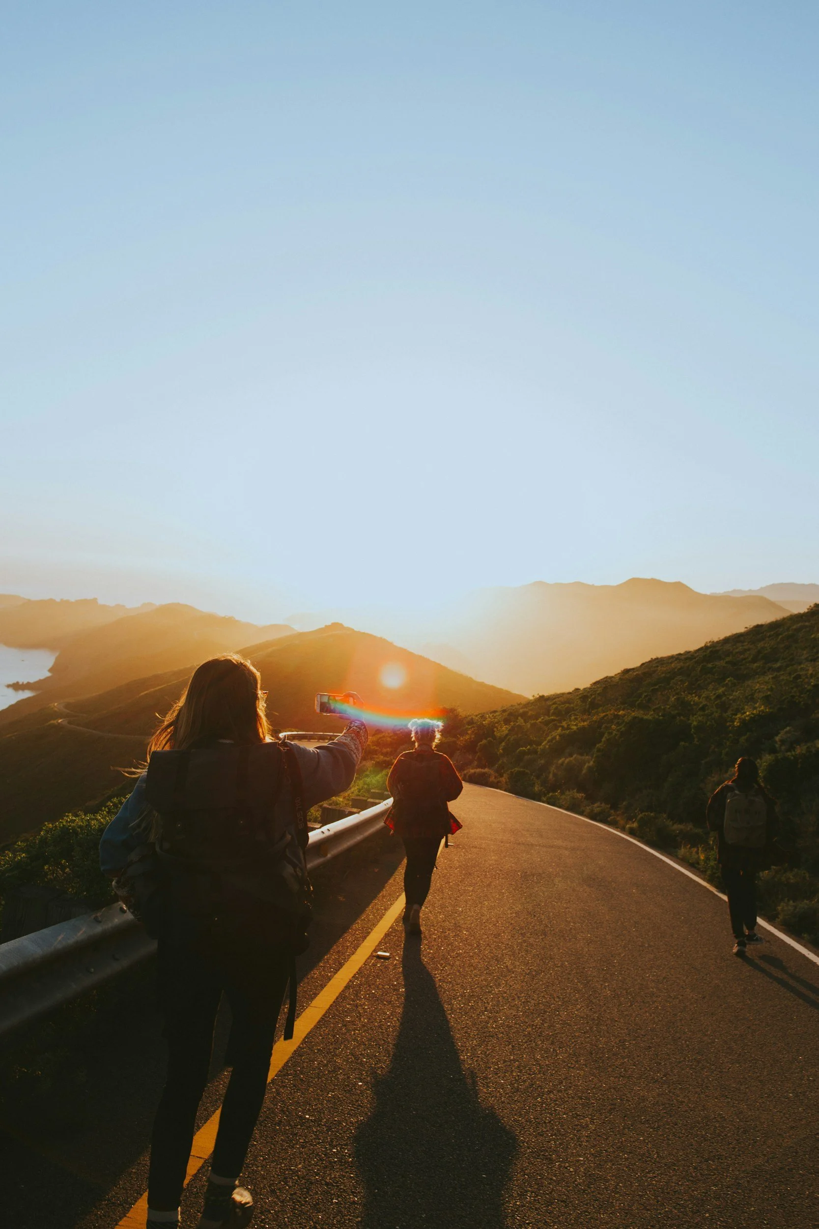 Three hikers walking on a winding mountain road at sunset, with one person taking a photo of the sunset.