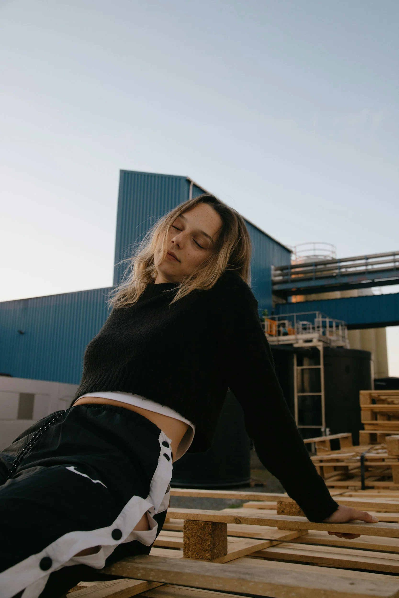 Young woman in black cropped sweater and black and white pants sitting on wooden pallets outdoors at an industrial site with blue building and sky in the background.