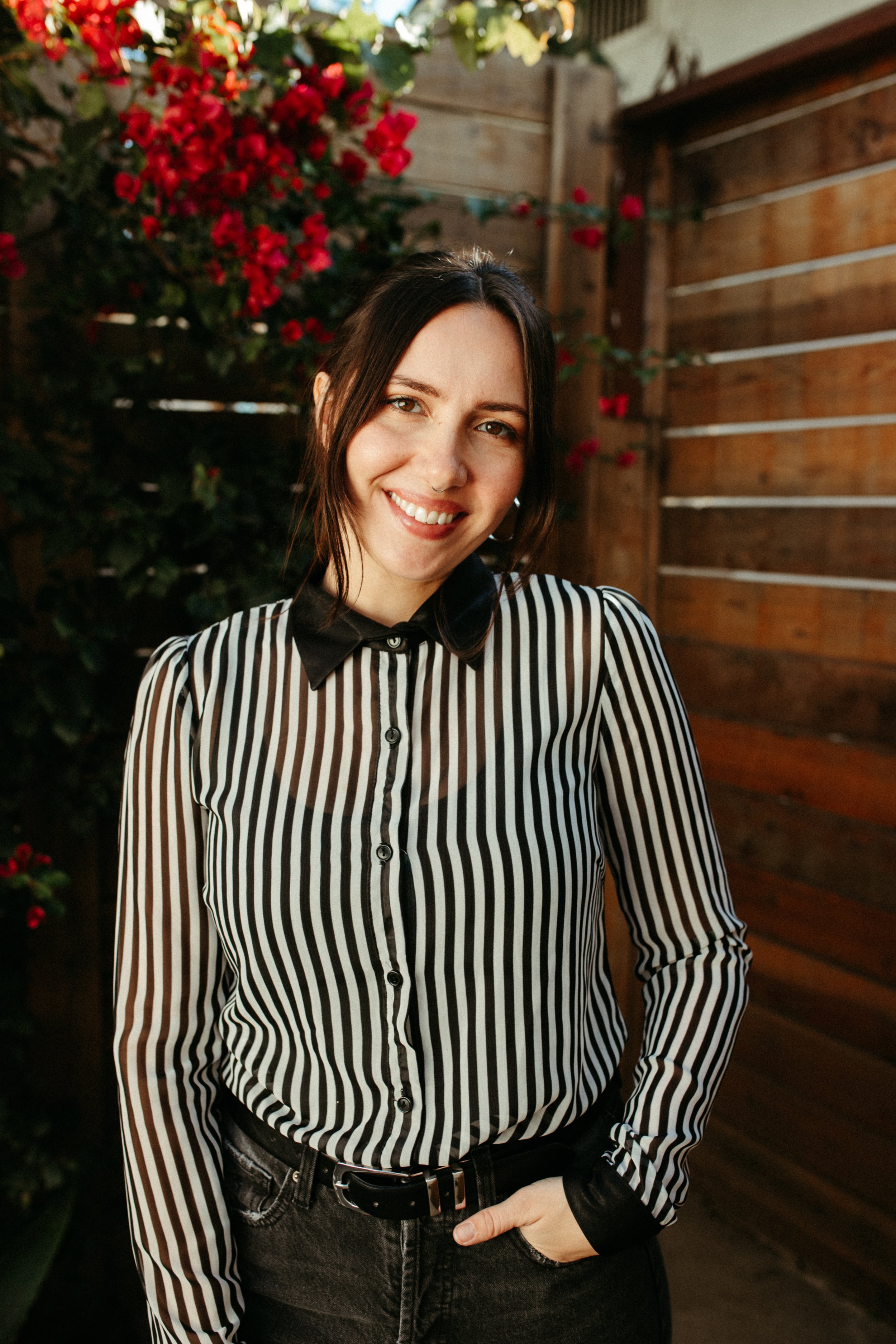 A young woman with dark hair smiling outdoors, wearing a black and white striped shirt and black pants, standing in front of a wooden fence and pink flowering bushes.