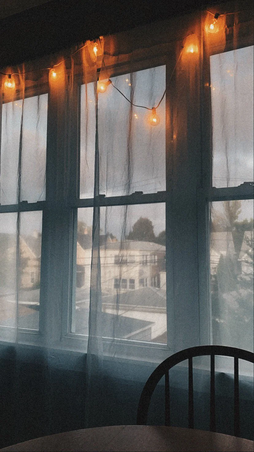 A window with sheer curtains, sunset outside, and warm string lights hanging above.