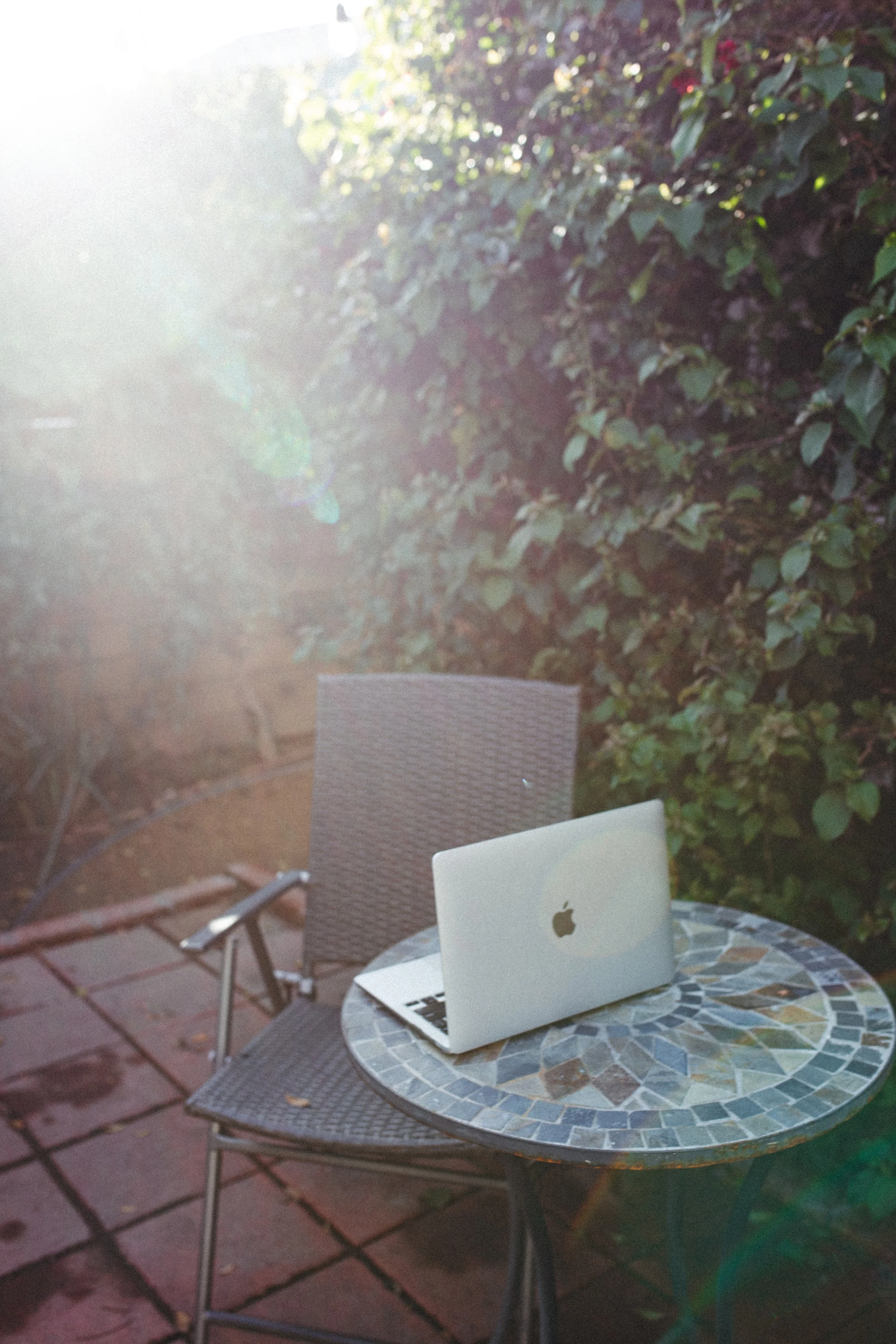 An outdoor patio scene with a round mosaic table and a chair, featuring a closed silver Apple MacBook on the table, surrounded by green leafy bushes in sunlight.