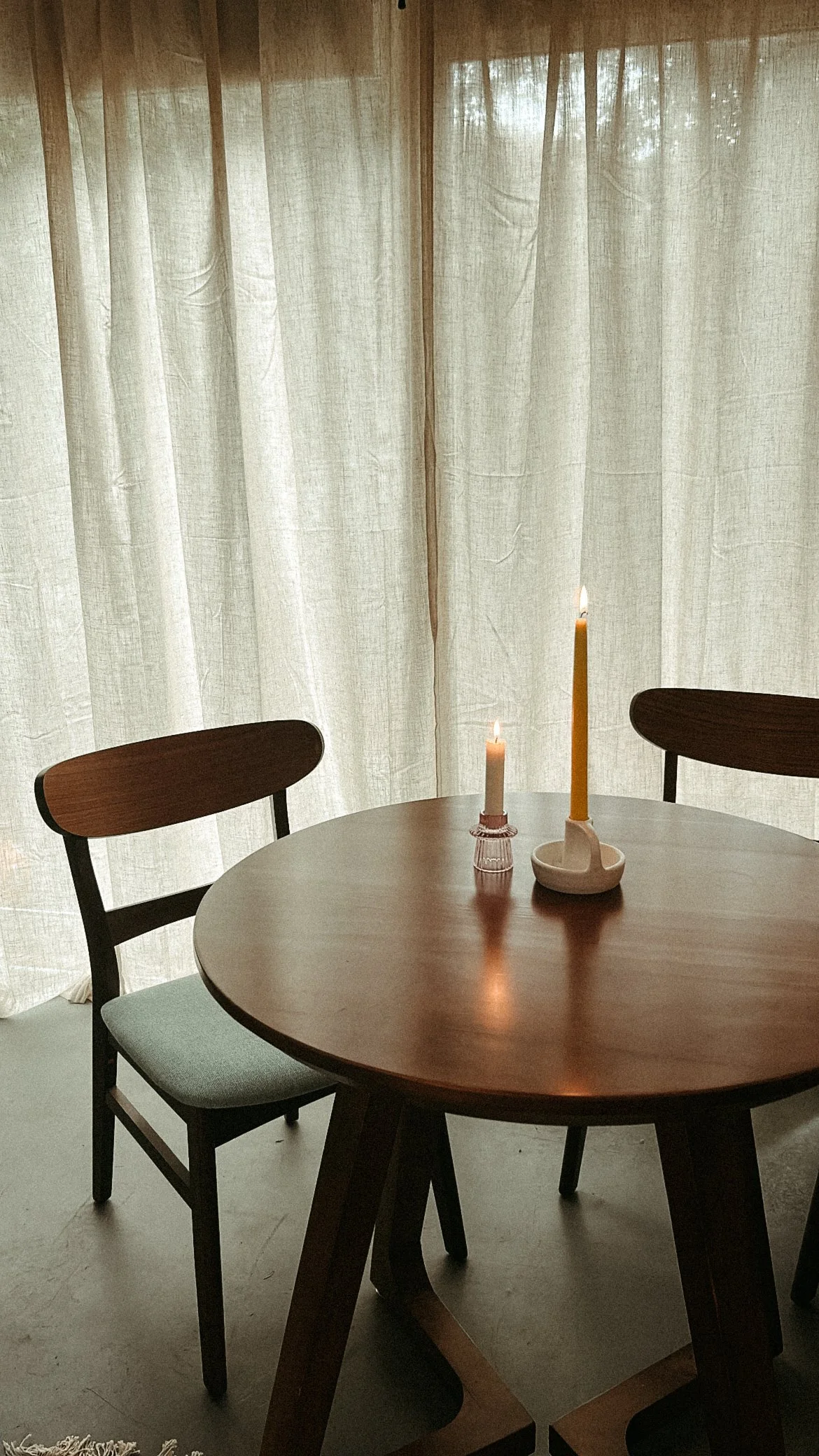 A wooden round dining table with two chairs, decorated with two lit candles in candle holders, set against a backdrop of light curtains.