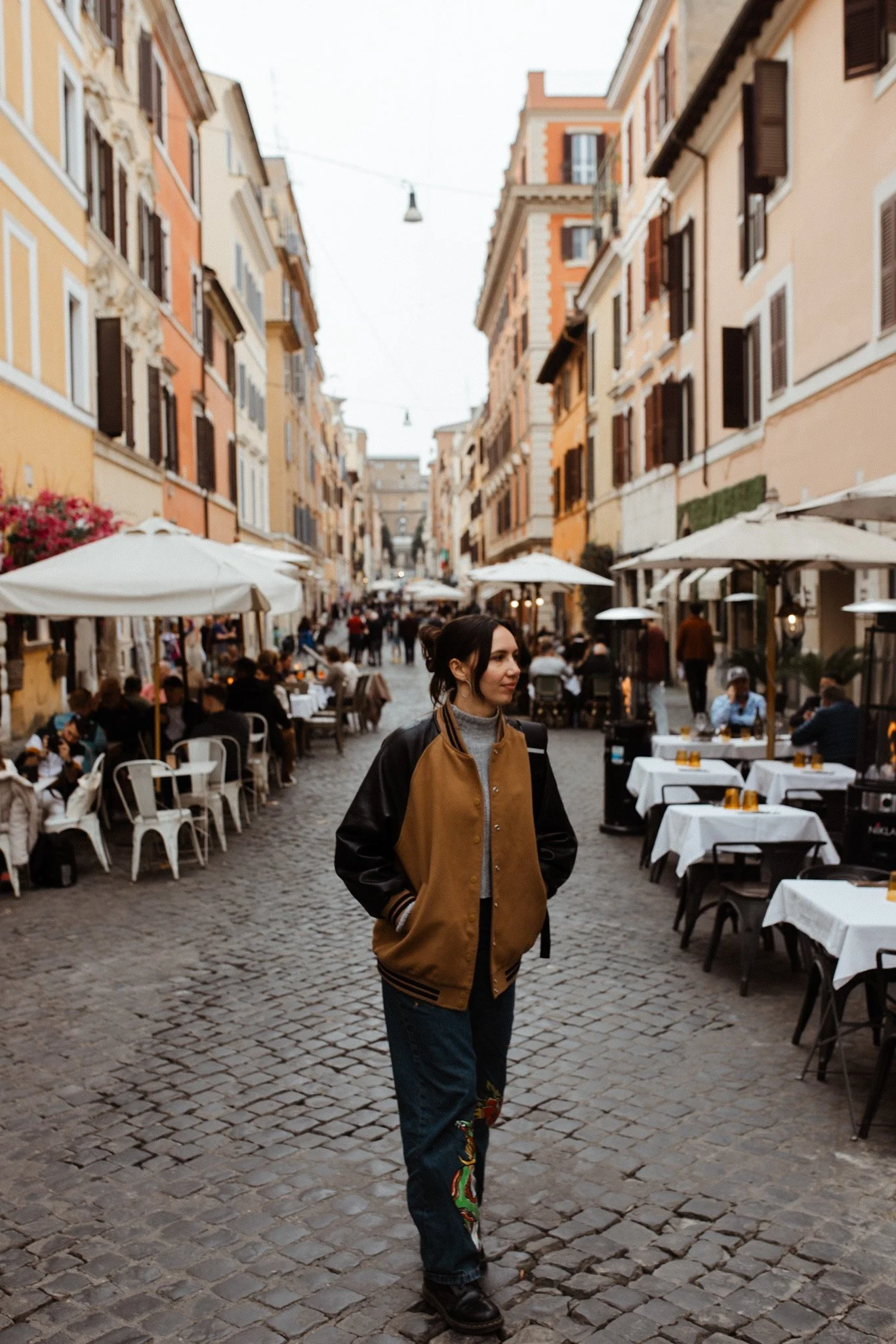 A woman wearing a brown and black varsity jacket and jeans with embroidery walks along a cobblestone street lined with outdoor cafes and colorful buildings.