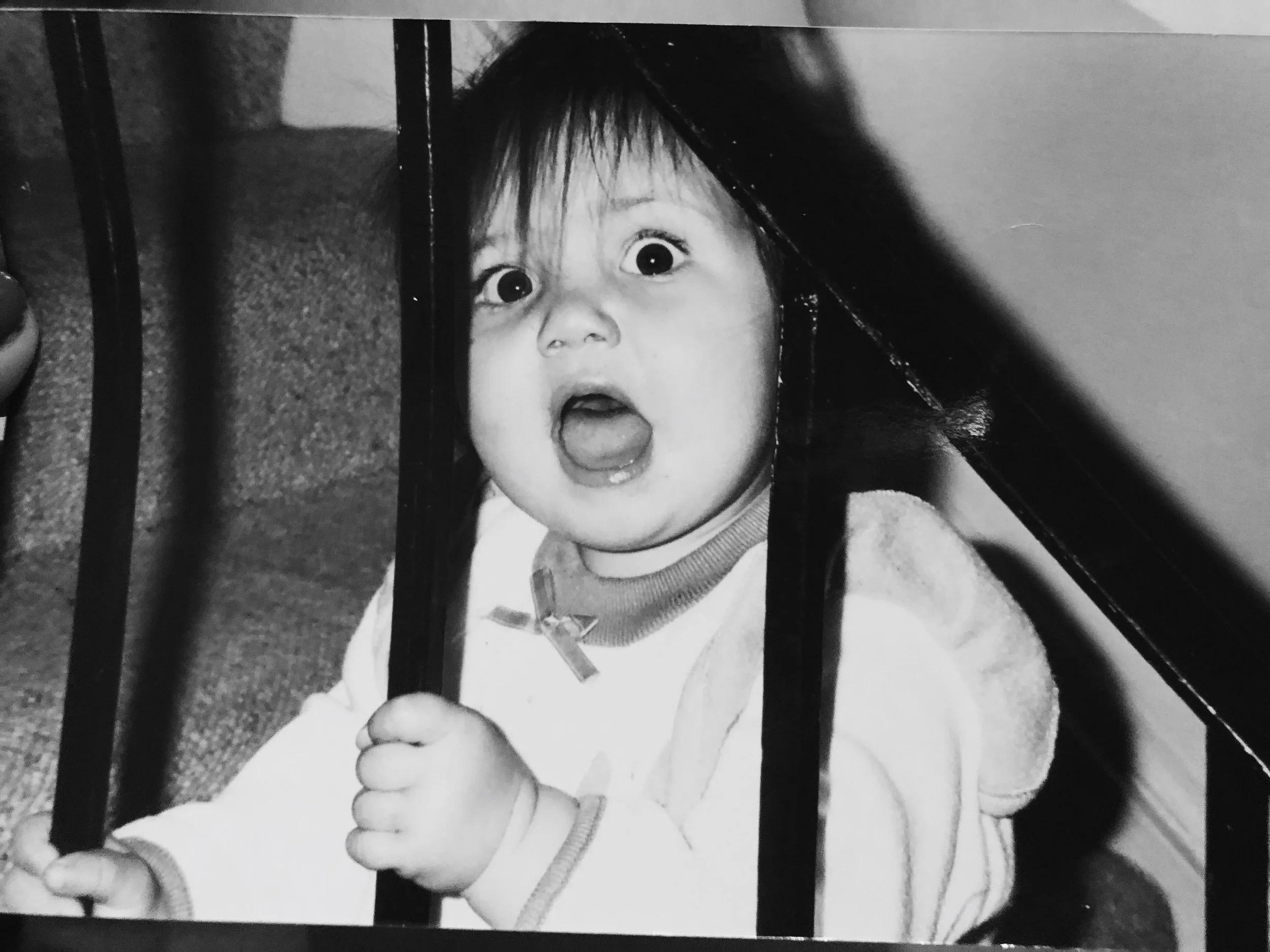 A young child with an expressive face, mouth open and eyes wide, holding onto a metal bar, possibly a railing or part of a piece of furniture.